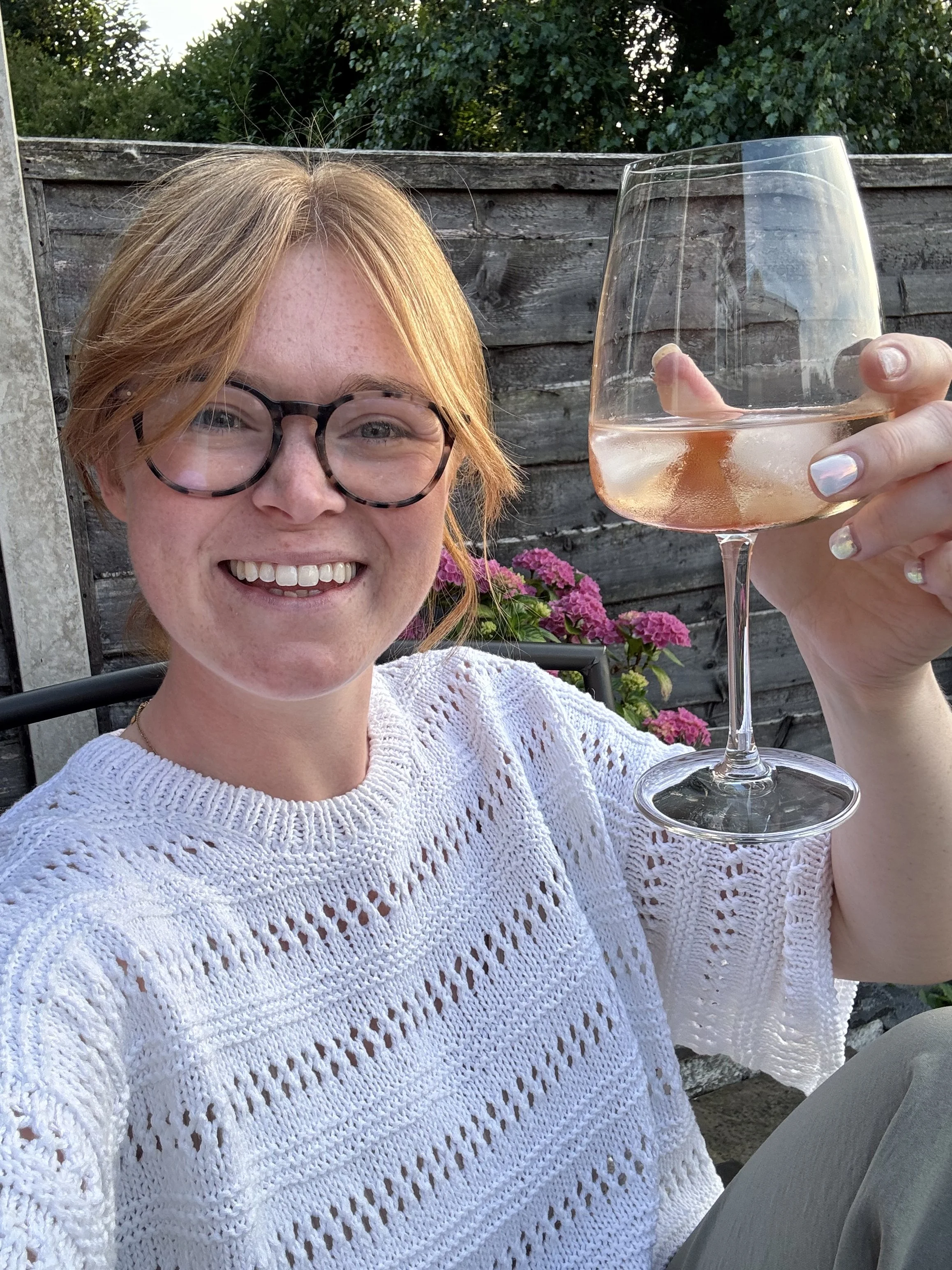 A woman (Lizzie who owns Hive Knits) with red hair, glasses, and a white knitted top (Lacey loop tee by the designer other loops) smiling and holding a glass of rosé wine outdoors, with a wooden fence and pink flowers in the background.