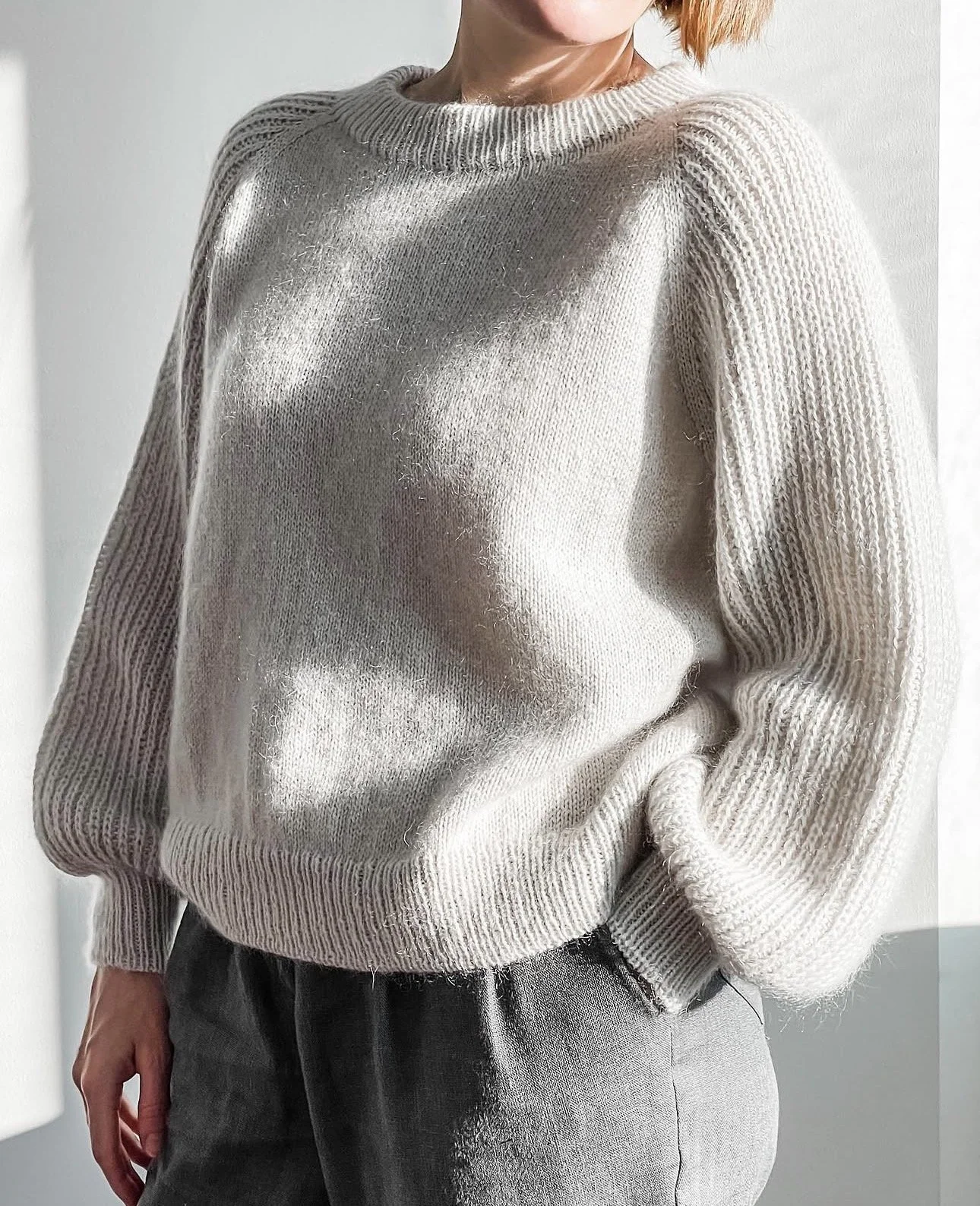 The First Sweater, a knitting pattern by Hive Knits. A person wearing a grey-cream-colored knit sweater with a crew-neck ribbed collar and textured Half Fisherman's Rib sleeves, paired with dark pants.