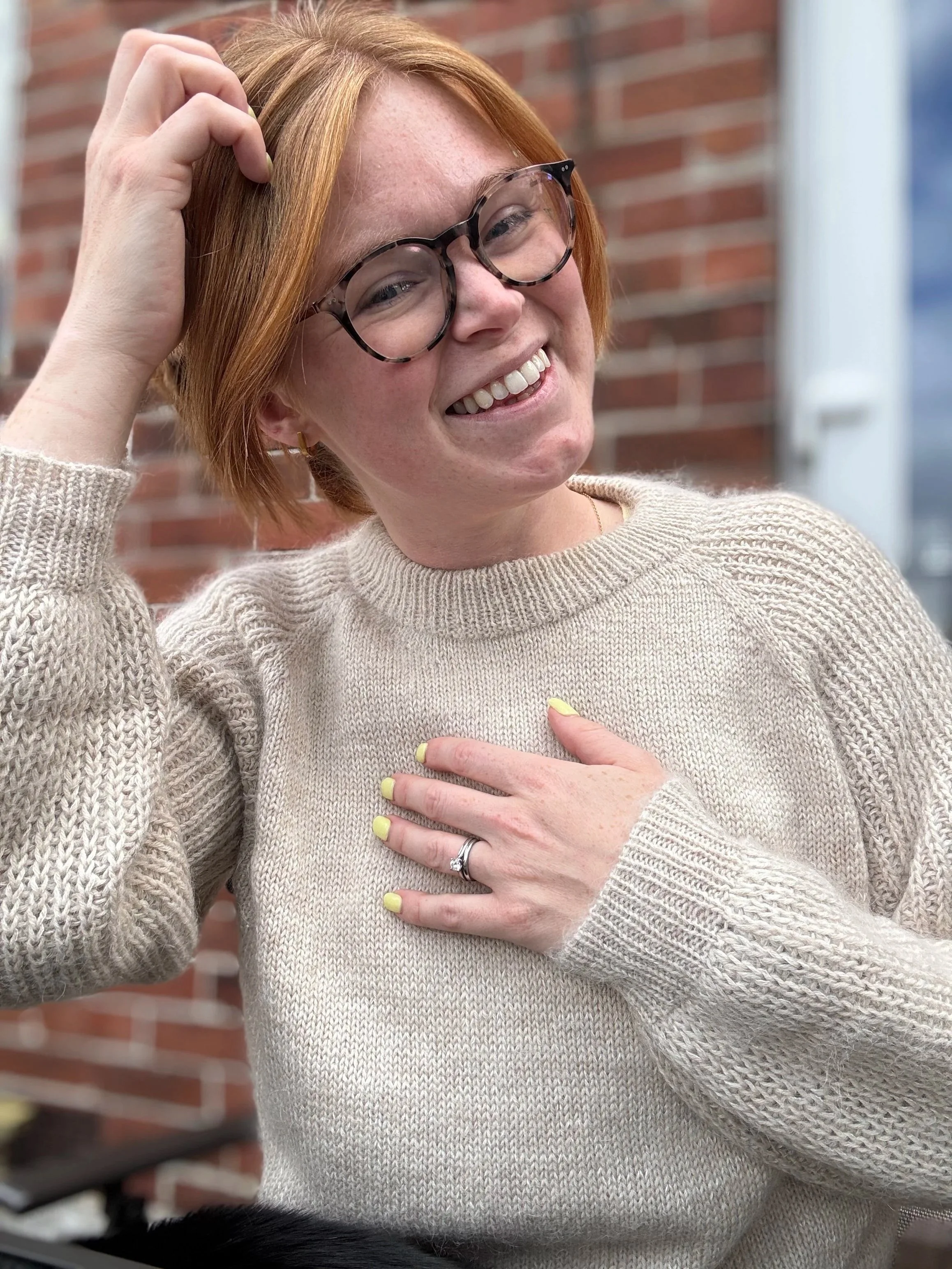 A woman with red hair and glasses smiling, wearing a beige sweater, touching her chest with her left hand which has a wedding ring, standing outside in front of a brick wall.