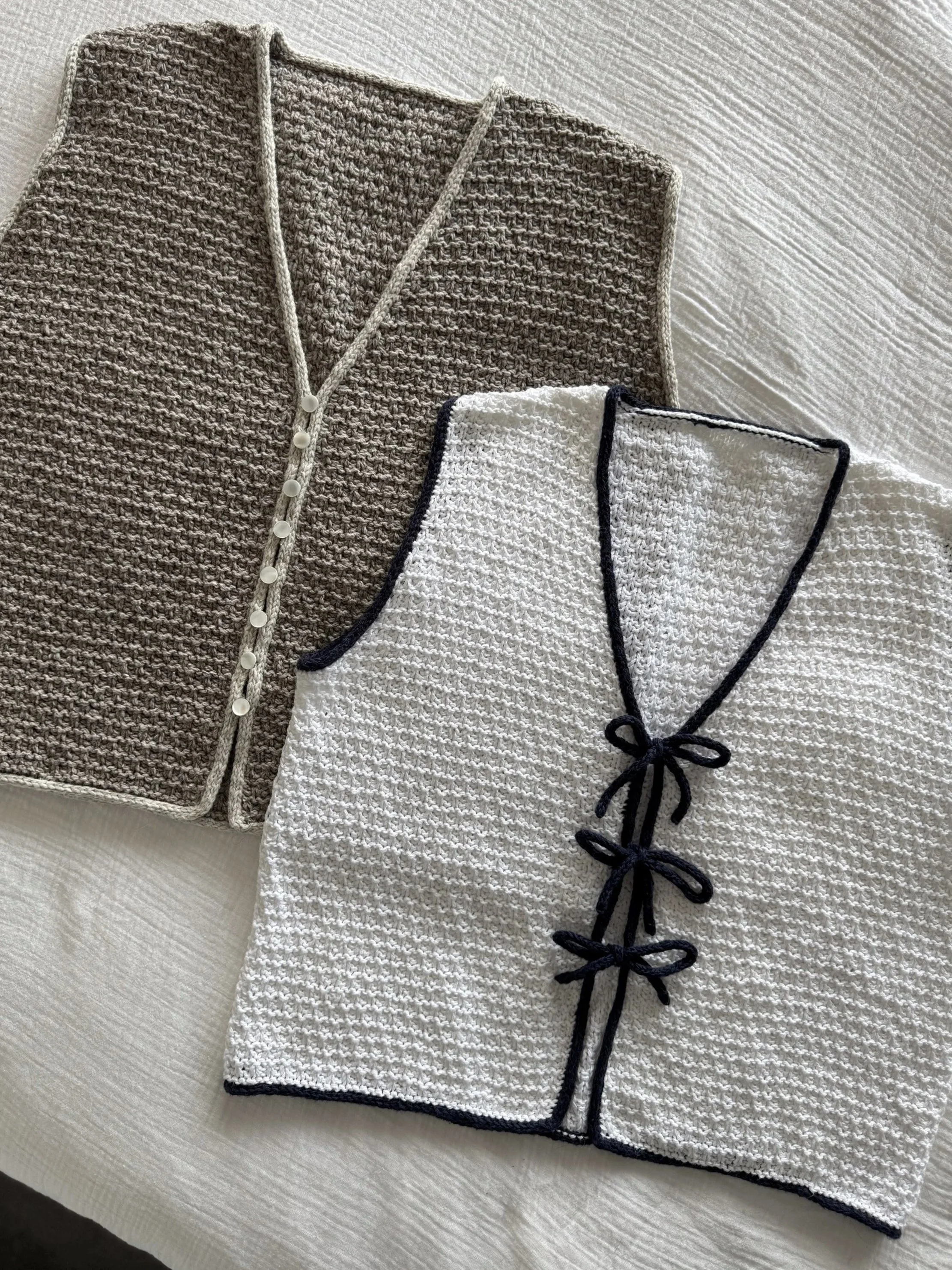 Tao Vest, a knitting pattern by Hive Knits. Two knitted v-neck vests, one in grey-brown with white buttons, and the other in white with navy i-cord trim and navy I-cord ties, laid on a white textured surface.