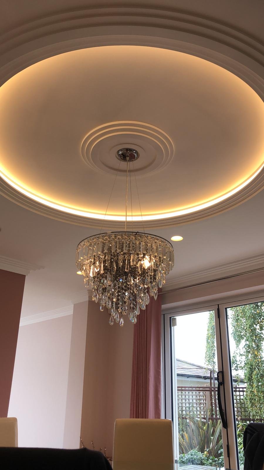 Elegant ceiling light fixture hanging from a decorative chandelier with crystal accents in a modern dining room with large windows and outdoor view.