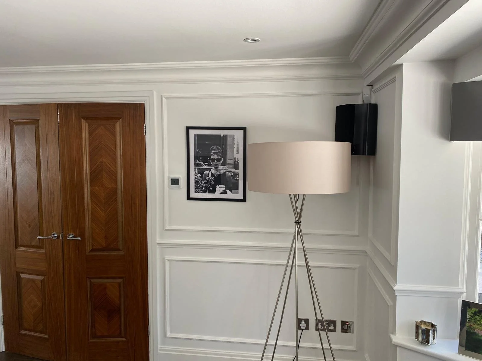 Interior of a room with a wooden door, a framed black and white photograph of a woman, a tall floor lamp with a beige shade, and a small TV mounted on the wall.