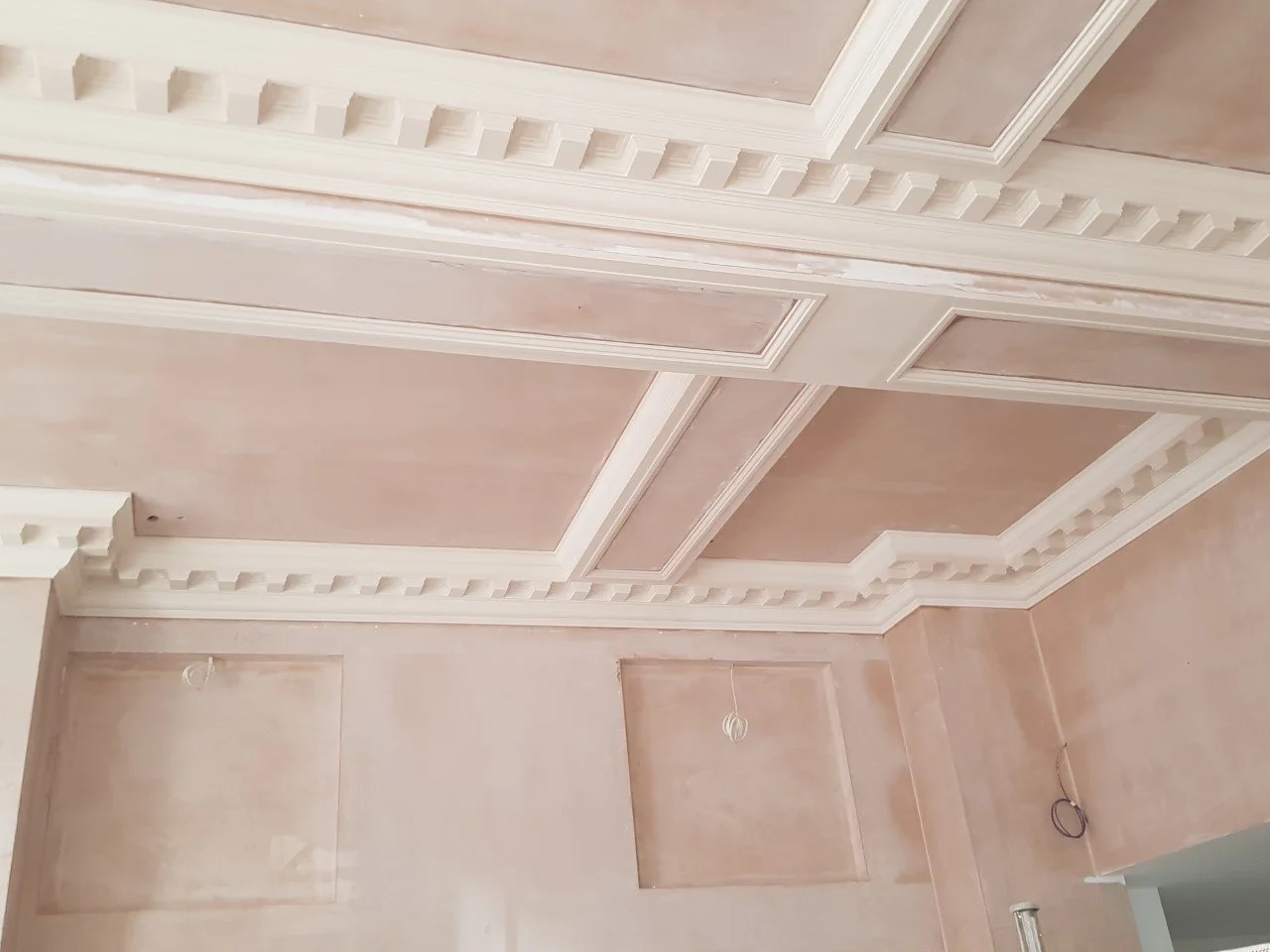 Room with decorative ceiling molding and recessed wall panels under construction, electrical wires hanging from the ceiling.