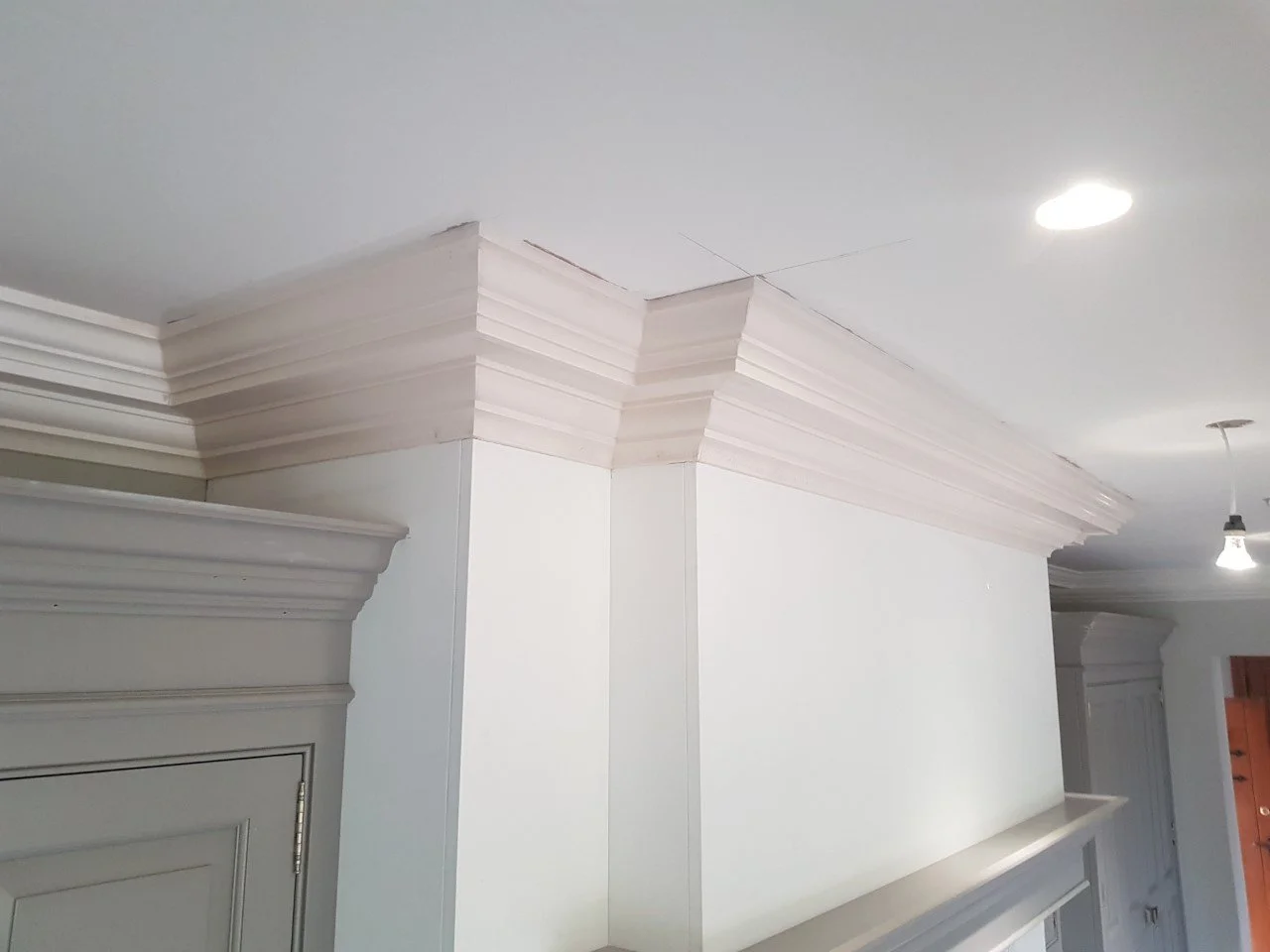 Interior ceiling corner with decorative crown molding and white walls, illuminated by ceiling lights.
