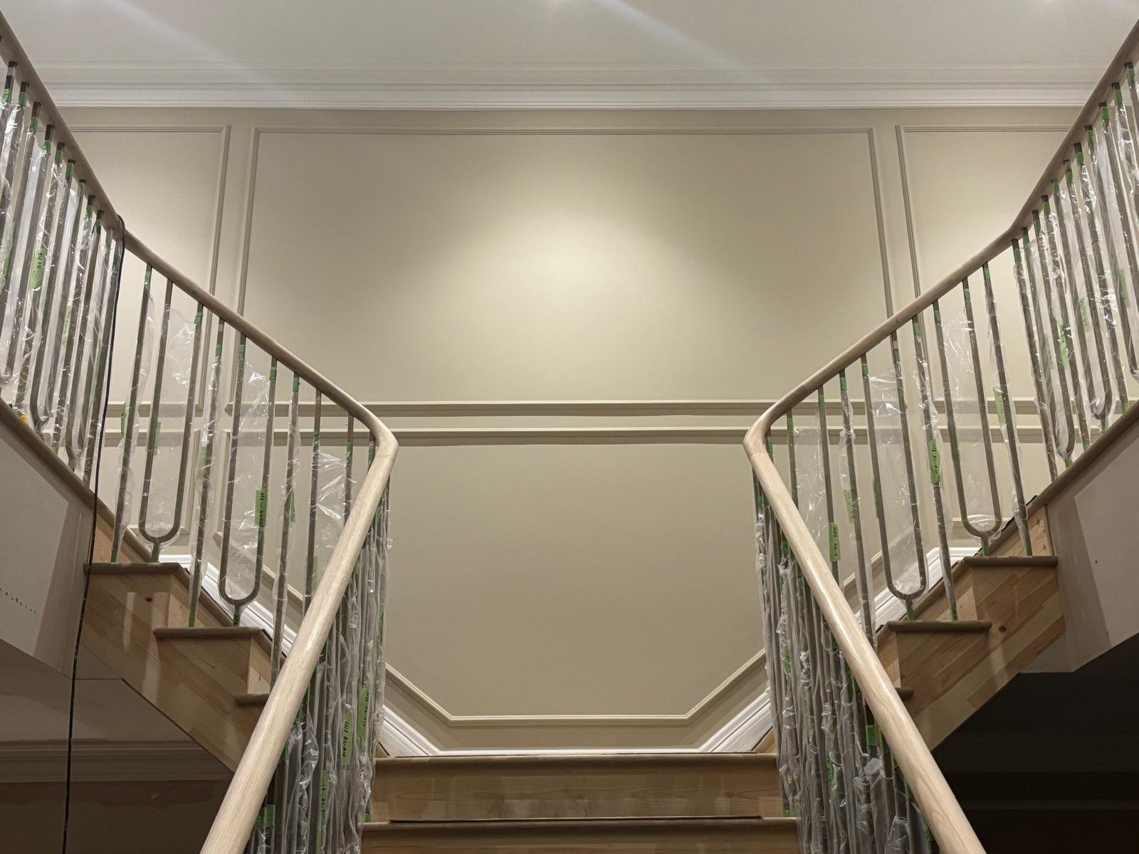 View of a staircase with new handrails and protective plastic wrapping, looking up towards a plain beige wall and ceiling.