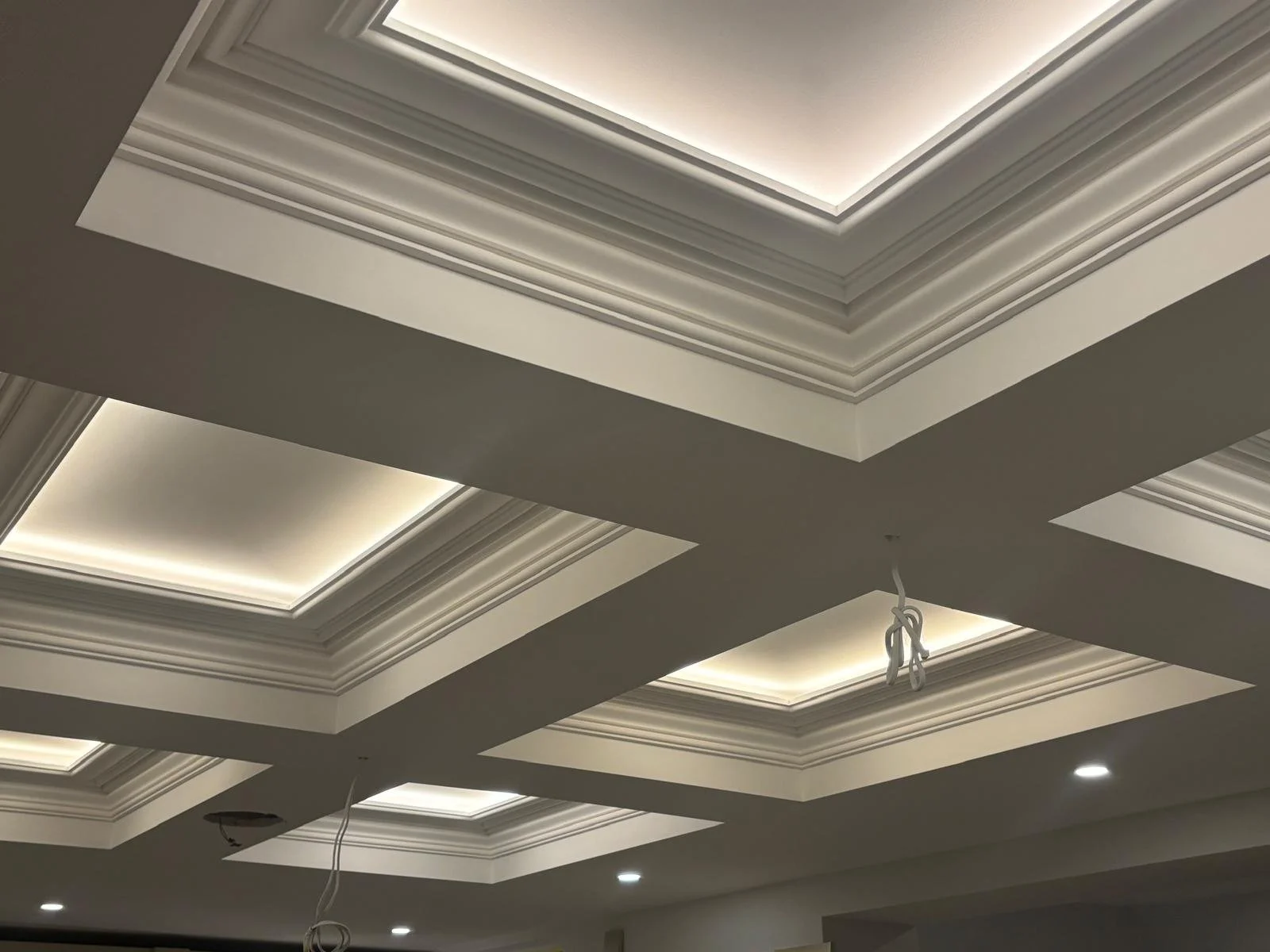Ceiling with decorative layered molding and built-in lighting fixtures, some wires hanging down, and recessed lights in the background.