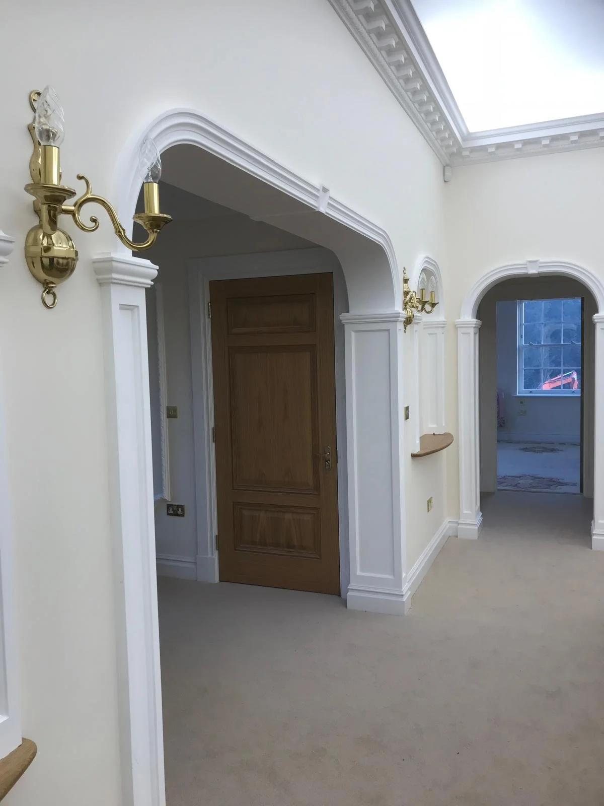 Interior of a house hallway with arched doorways, cream-colored walls, wooden doors, brass wall sconces, and a skylight ceiling.