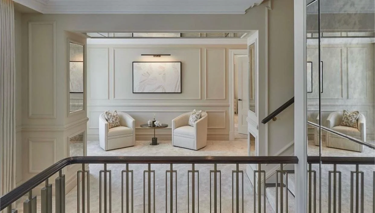 Interior view of a residential space with a sitting area featuring two white armchairs, a small round table between them, a framed artwork on the wall, and mirrored wall panels reflecting the space.