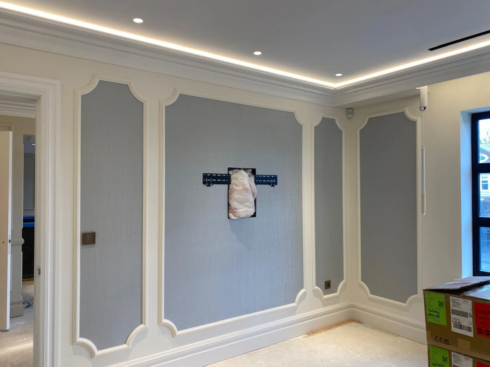Interior view of a room under renovation, with decorative wall paneling and a mounted TV bracket wrapped in plastic in the center. There is crown molding, recessed lighting in the ceiling, and a window on the right.