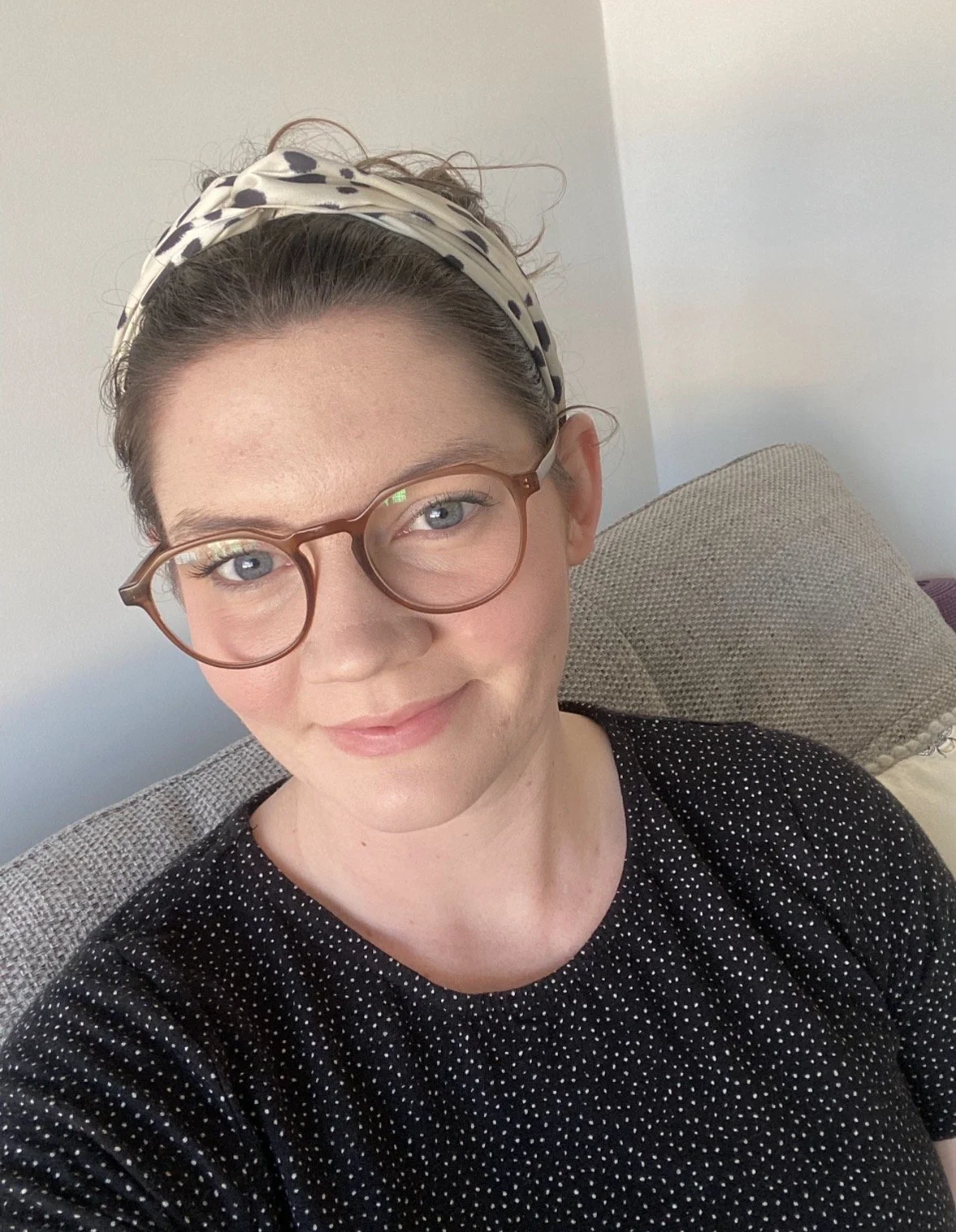A woman with short brown hair, glasses, and a polka-dot top sits on a gray couch with textured cushions in a room with light-colored walls, smiling at the camera.
