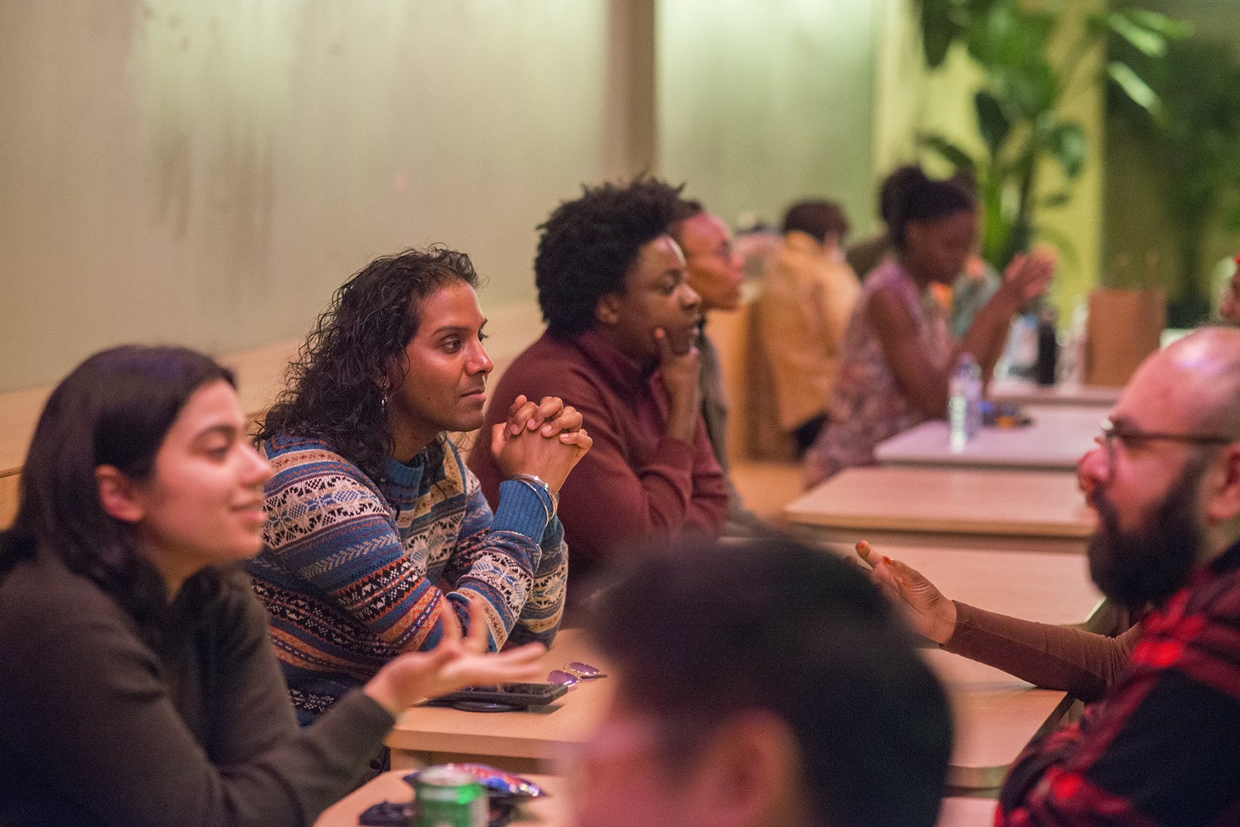 People seated at a long conference table, listening attentively, with some taking notes, in a room with green mural and potted plants in the background.