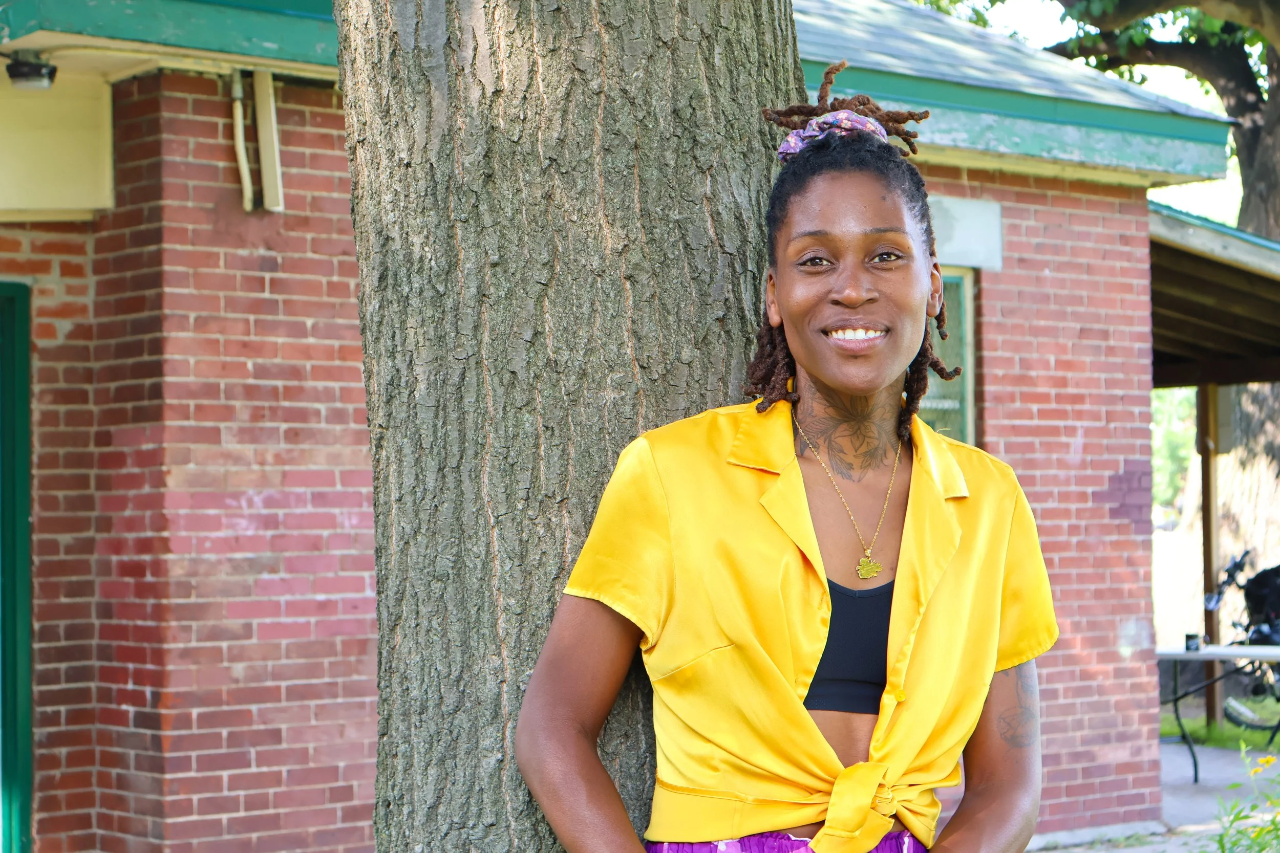 A woman with dreadlocks wearing a yellow shirt standing outdoors beside a tree with a brick building in the background.