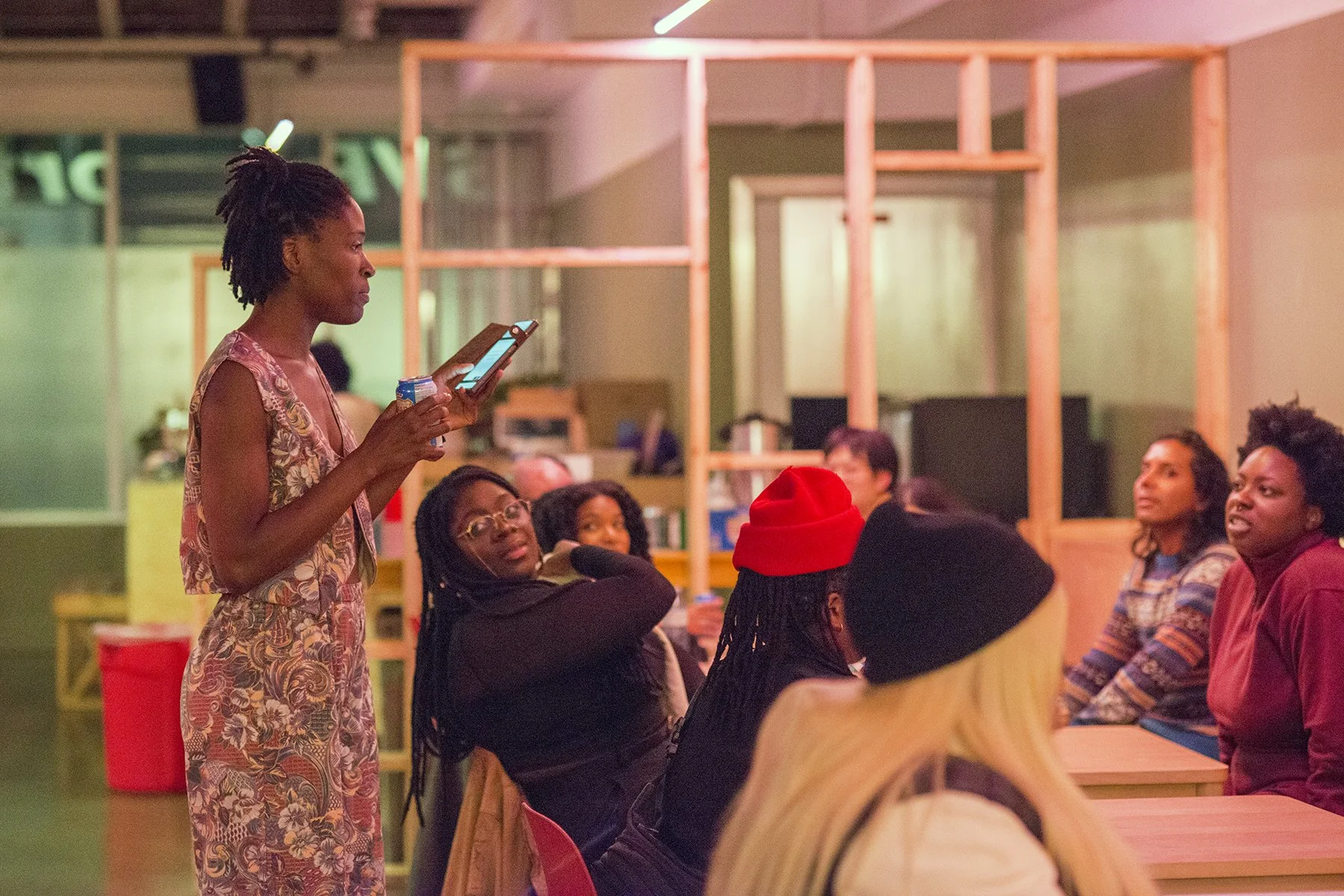 A woman standing and speaking to a group of people seated at a table in an indoor space with a wooden frame wall in the background.