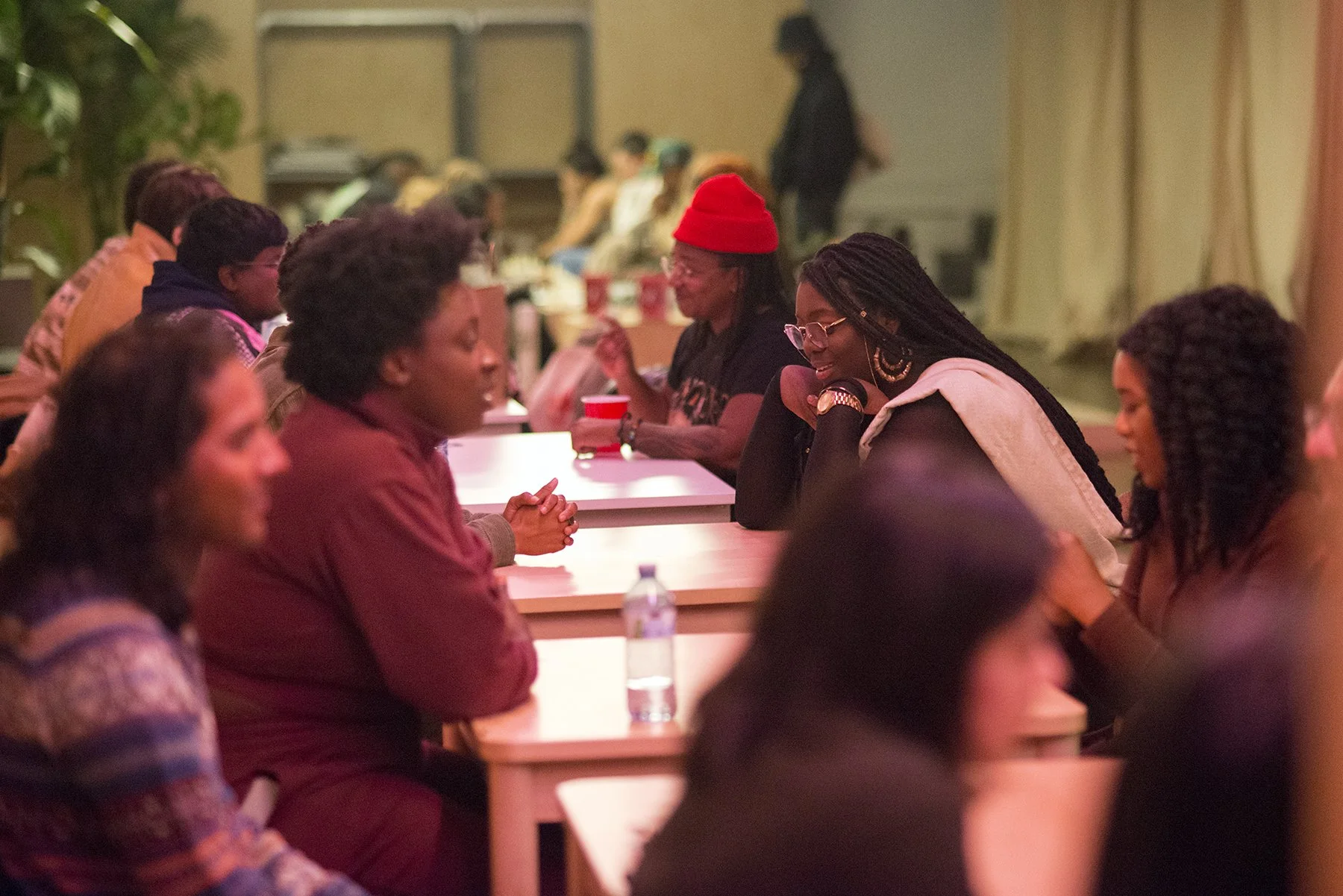 A group of people sitting at a long table in a dimly lit room, engaged in conversations. The focus is on two women in the center smiling and talking to each other, one wearing glasses and a light-colored shirt with a hoodie, and the other wearing a r