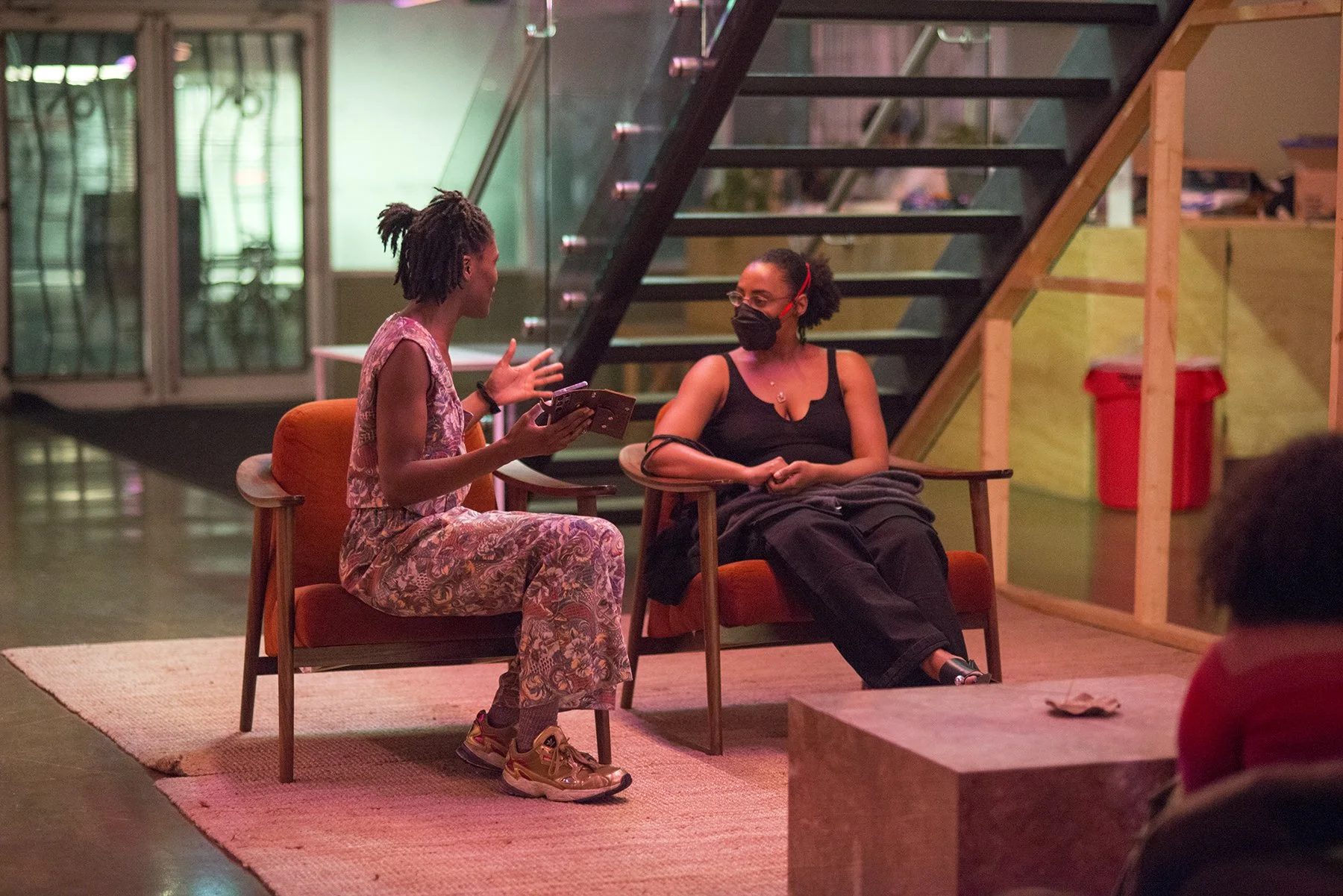 Two women seated in chairs having a conversation in a cozy indoor setting. One woman is wearing a floral dress and sneakers, holding a pamphlet, and gesturing with her hand. The other woman is dressed in black, wearing a face mask, and listening atte