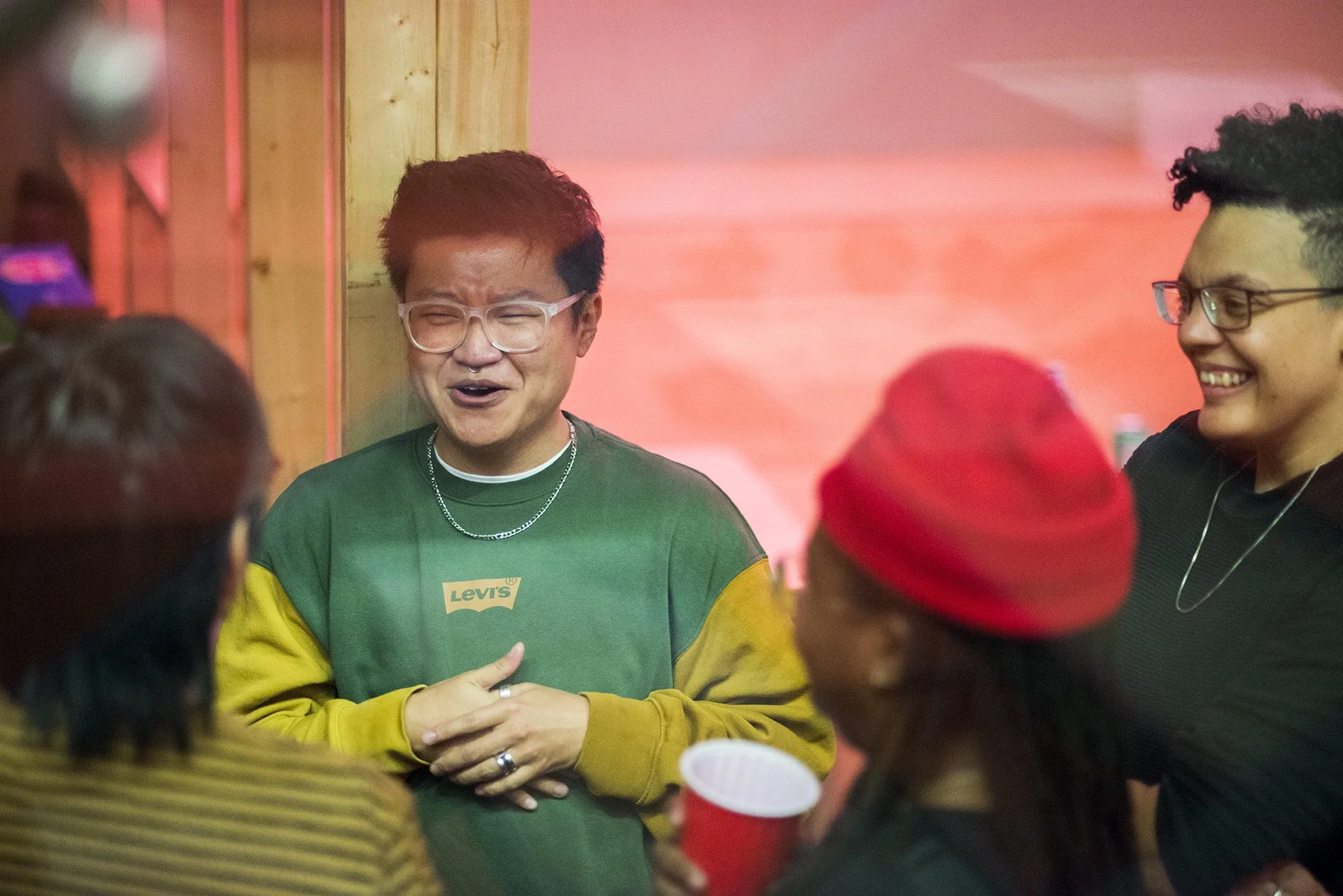 Group of people, two are smiling, engaging in conversation, standing indoors with wooden wall and pink lighting in the background.