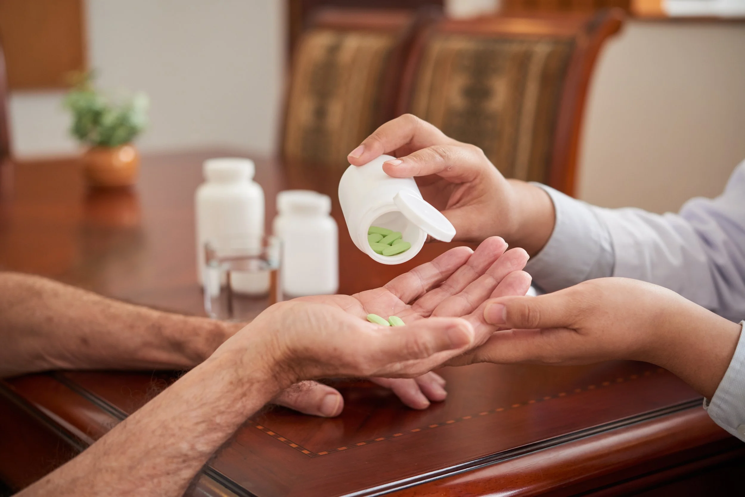 A person dispensing green pills from a white bottle into another person's hand, with a second bottle nearby, placed on a wooden table.