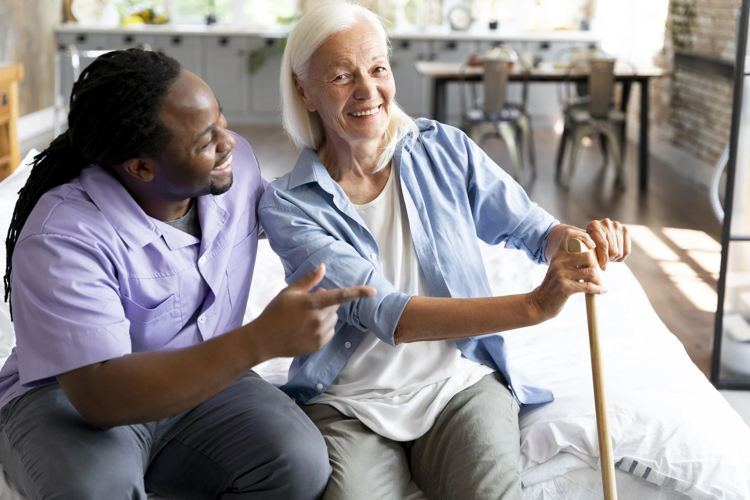 A smiling elderly woman in a blue shirt holding a cane, sitting next to a smiling man in a purple shirt in a bright, modern kitchen with a dining area in the background.