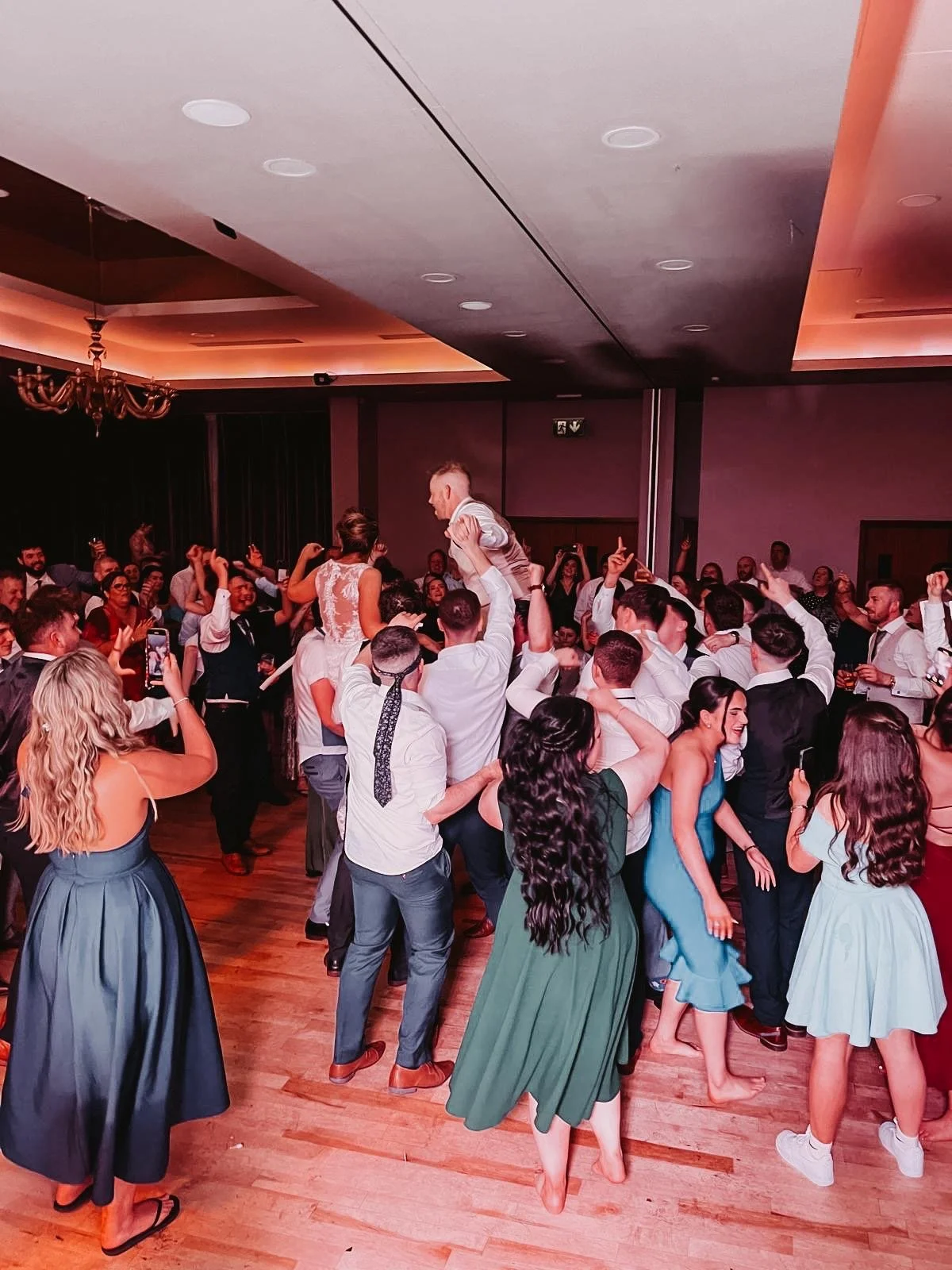 People dancing and celebrating at a wedding reception or party, with some guests taking photos, in a large indoor venue with warm lighting and a chandelier.
