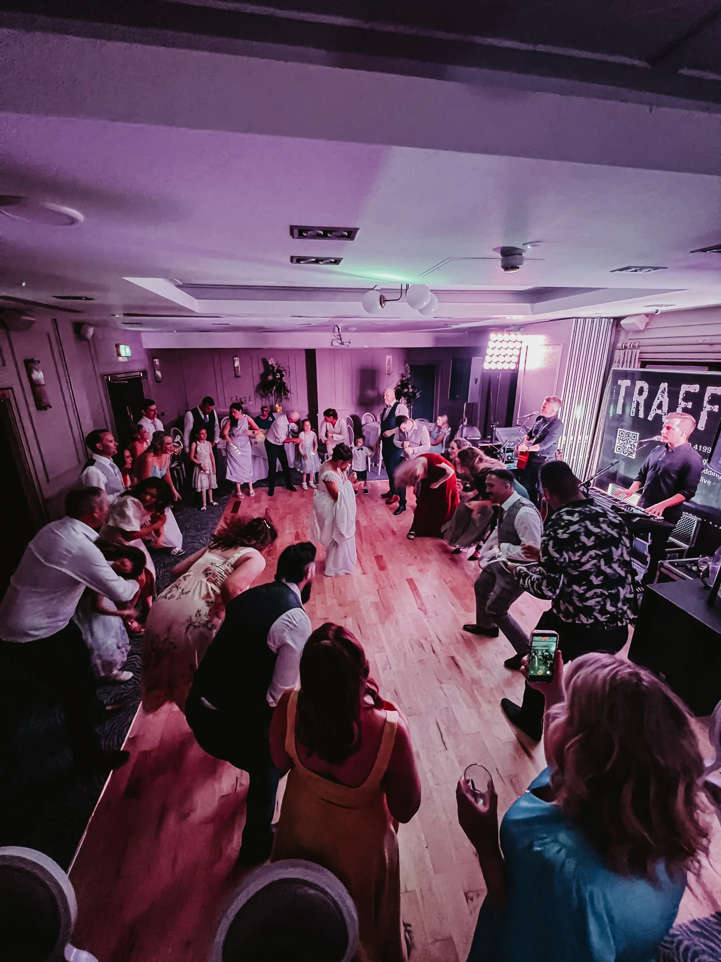 People gathered on a dance floor at a party with a band playing on stage, purple lighting, and decorations.