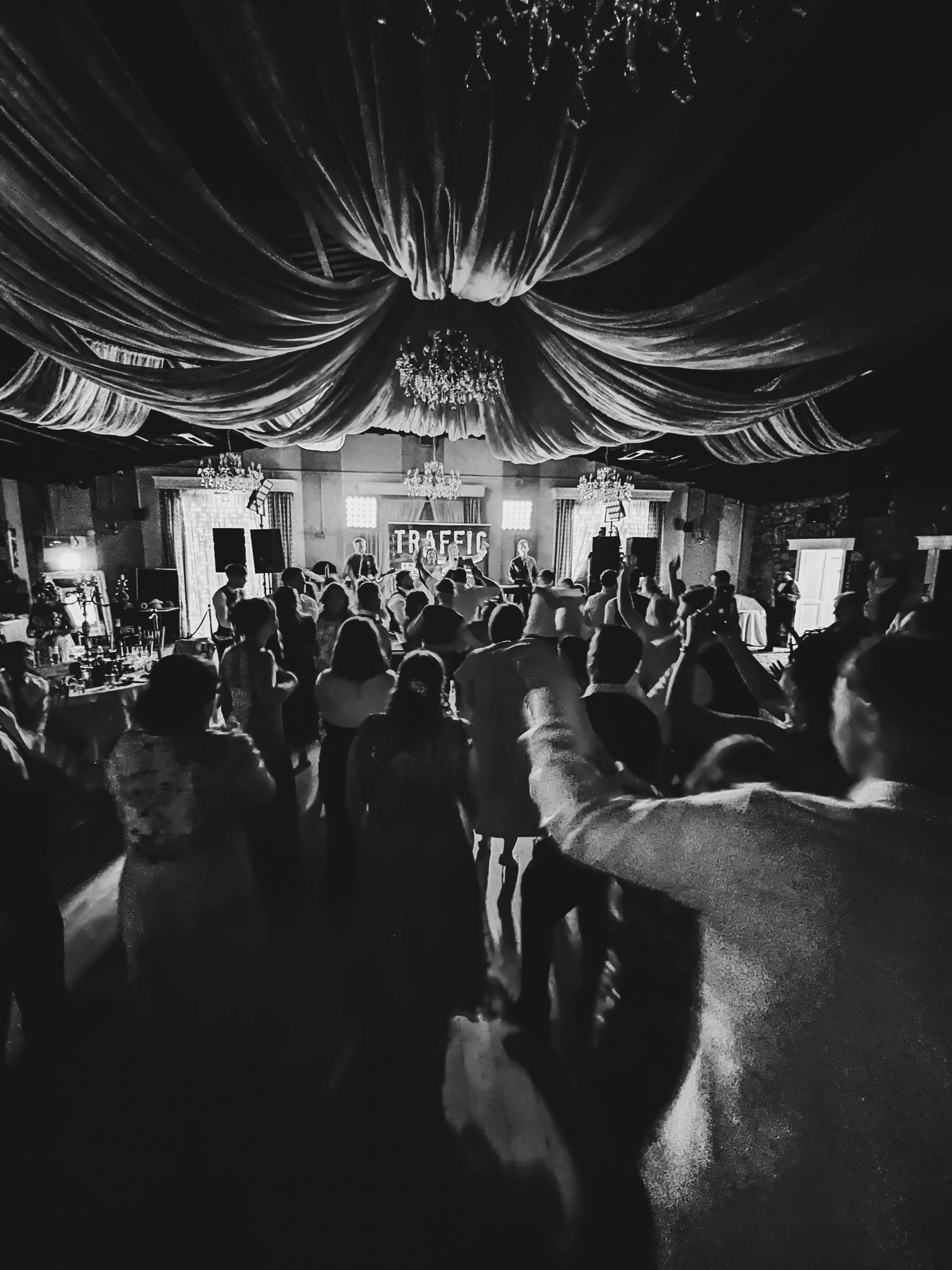 Black and white photo of a lively indoor event with a stage, a band performing, a crowd dancing and socializing, elegant drapery on the ceiling, and a sign with the word 'TRAFIC' in the background.
