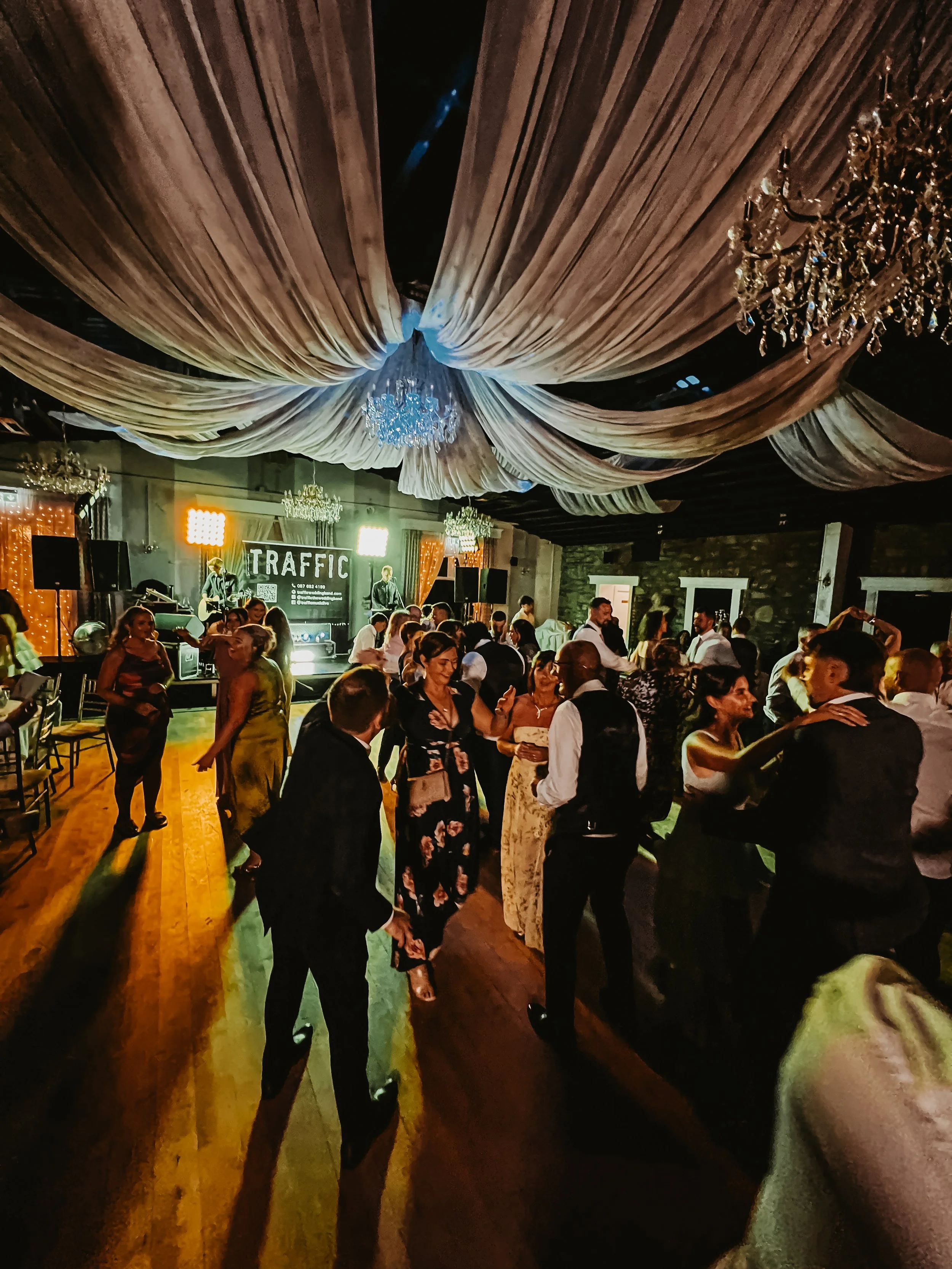 A lively Irish wedding with people dancing and socializing. The ceiling is decorated with large, flowing drapery and chandeliers. A live band performs on stage with a sign that says 'TRAFFIC'.