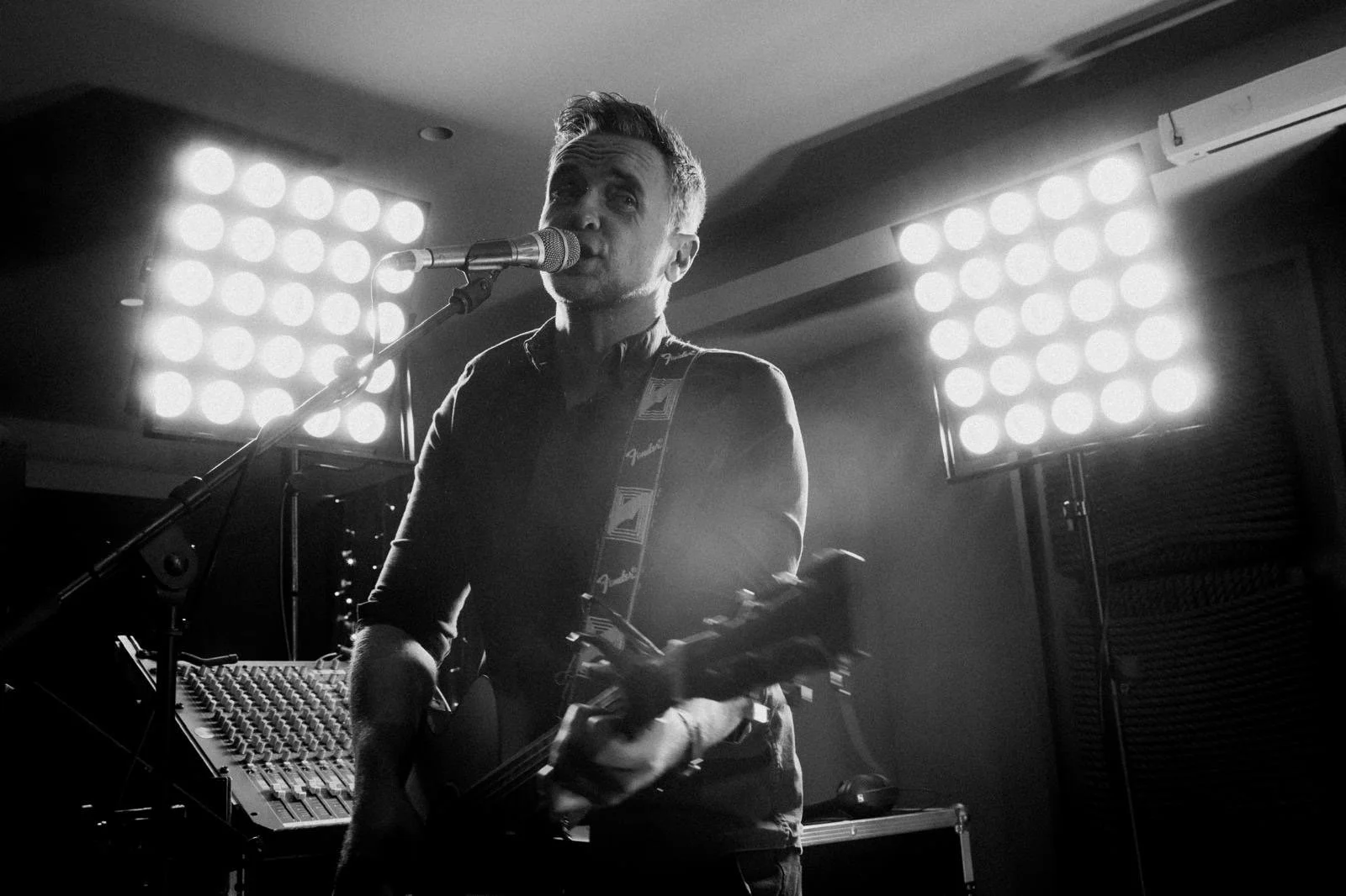 A black-and-white photo of Jimmy Fyfe singing into a microphone and playing an electric guitar at a wedding. Bright stage lights are visible behind him.