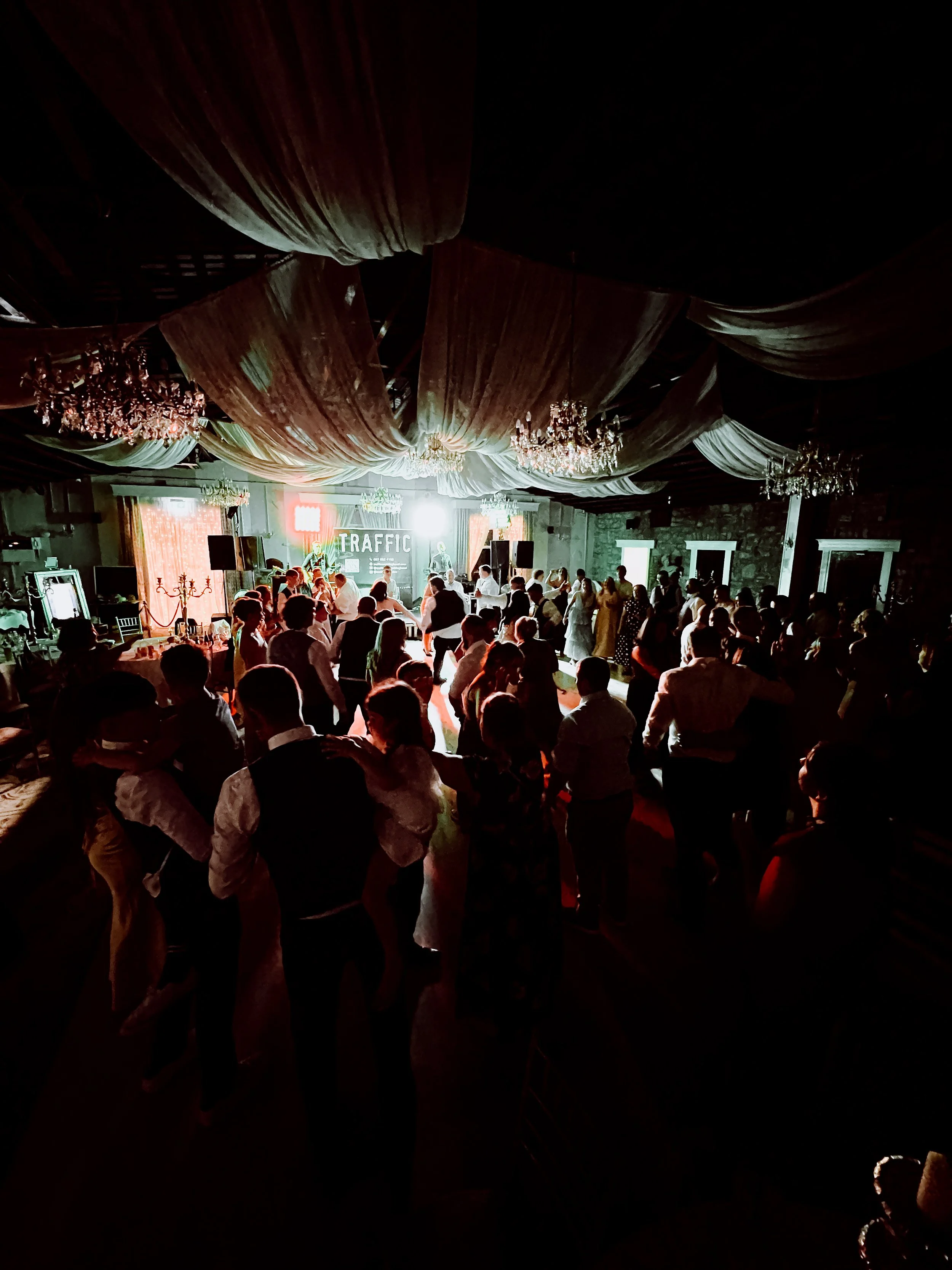 A crowded indoor party or event with people dancing and socializing, decorated with chandeliers and drapery on the ceiling, with a stage and bright lights in the background.