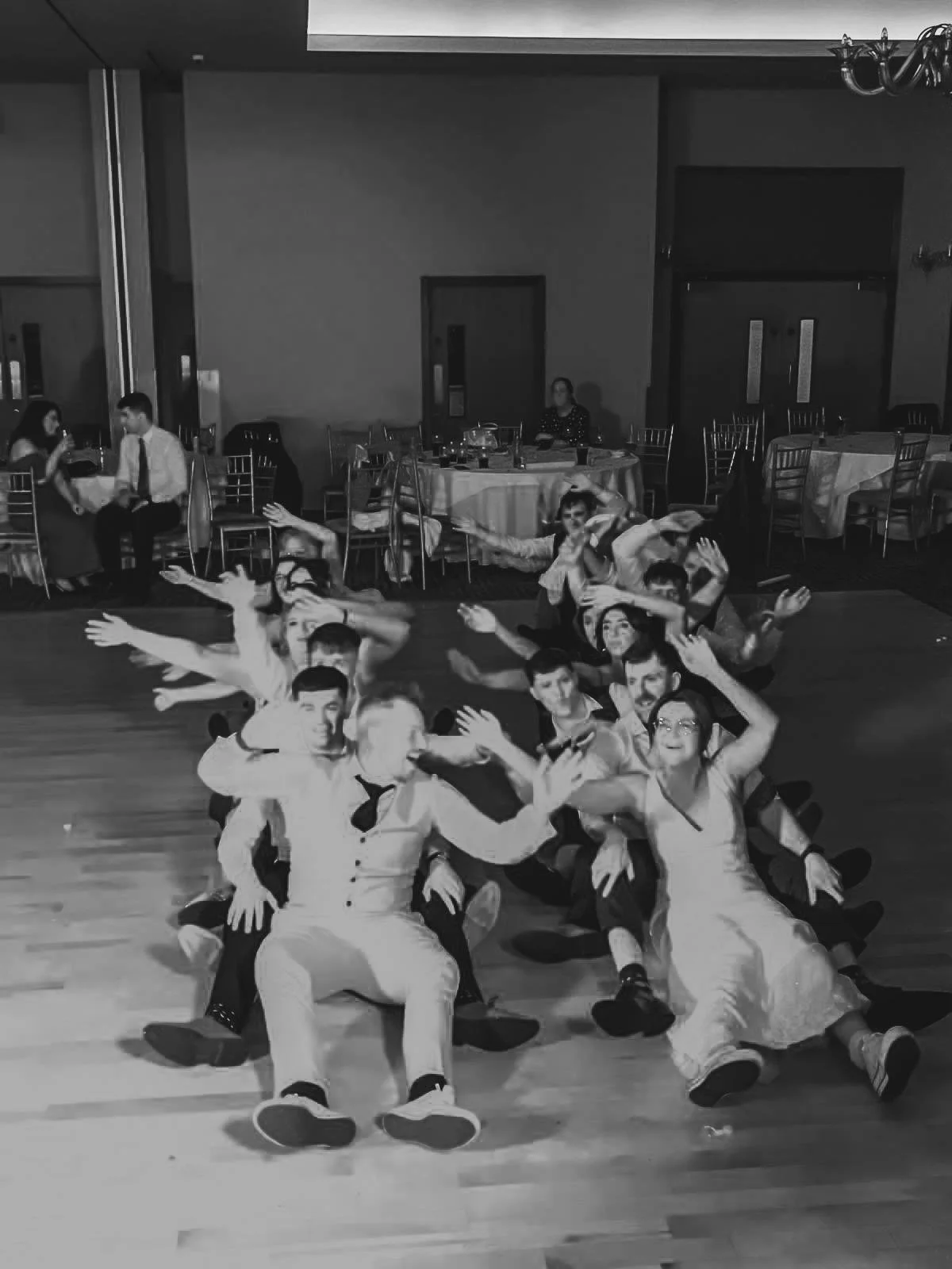 Group of people sitting on the floor in a line, smiling and posing with their arms outstretched, at an event or party in a banquet hall.