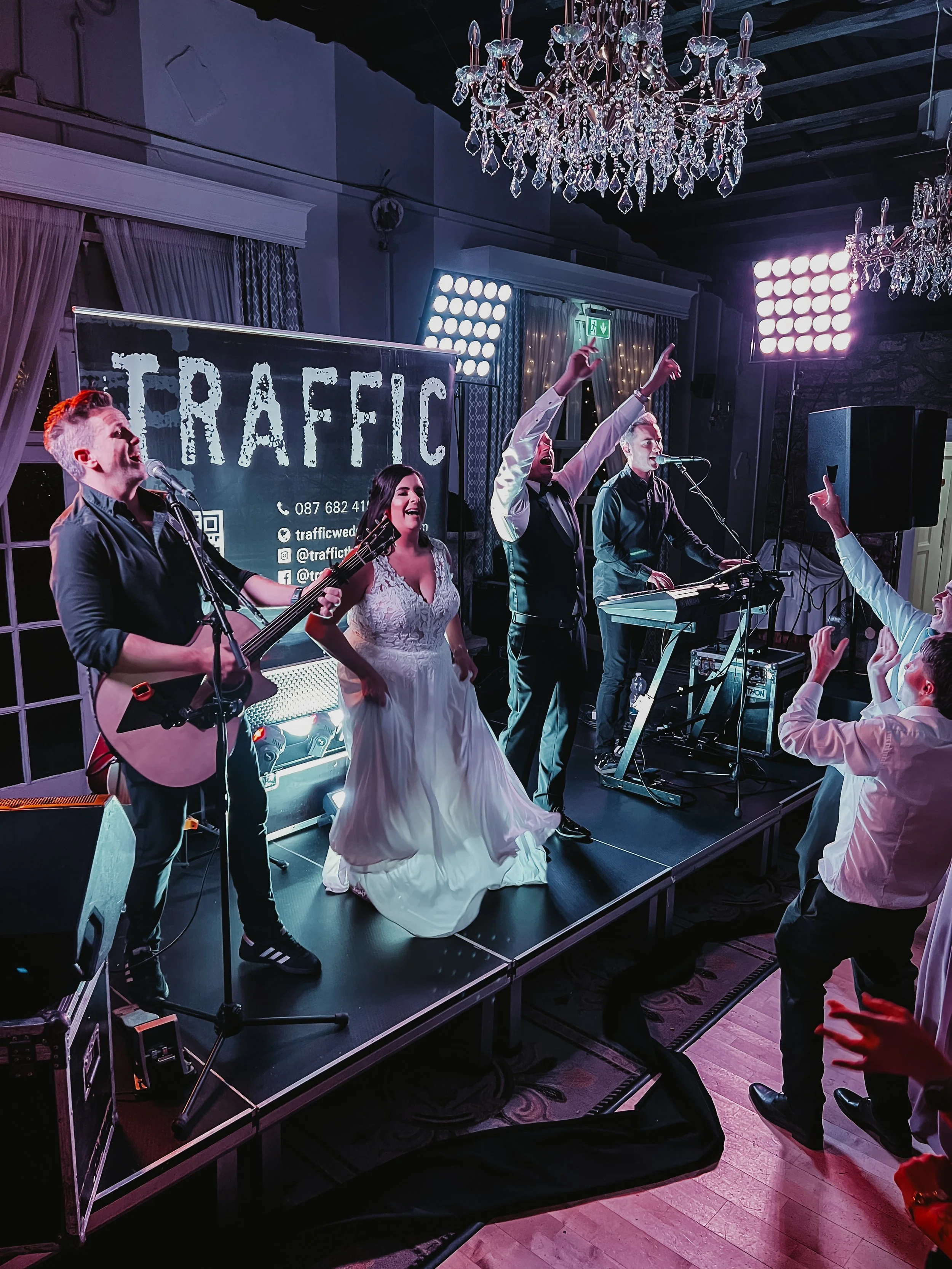 A musical performance on a stage with a band and a woman singing in a white dress, surrounded by singing and playing instruments. Audience members are visible in the foreground, and a large chandelier hangs above the stage with a sign reading 'TRAFFIC' behind the performers.