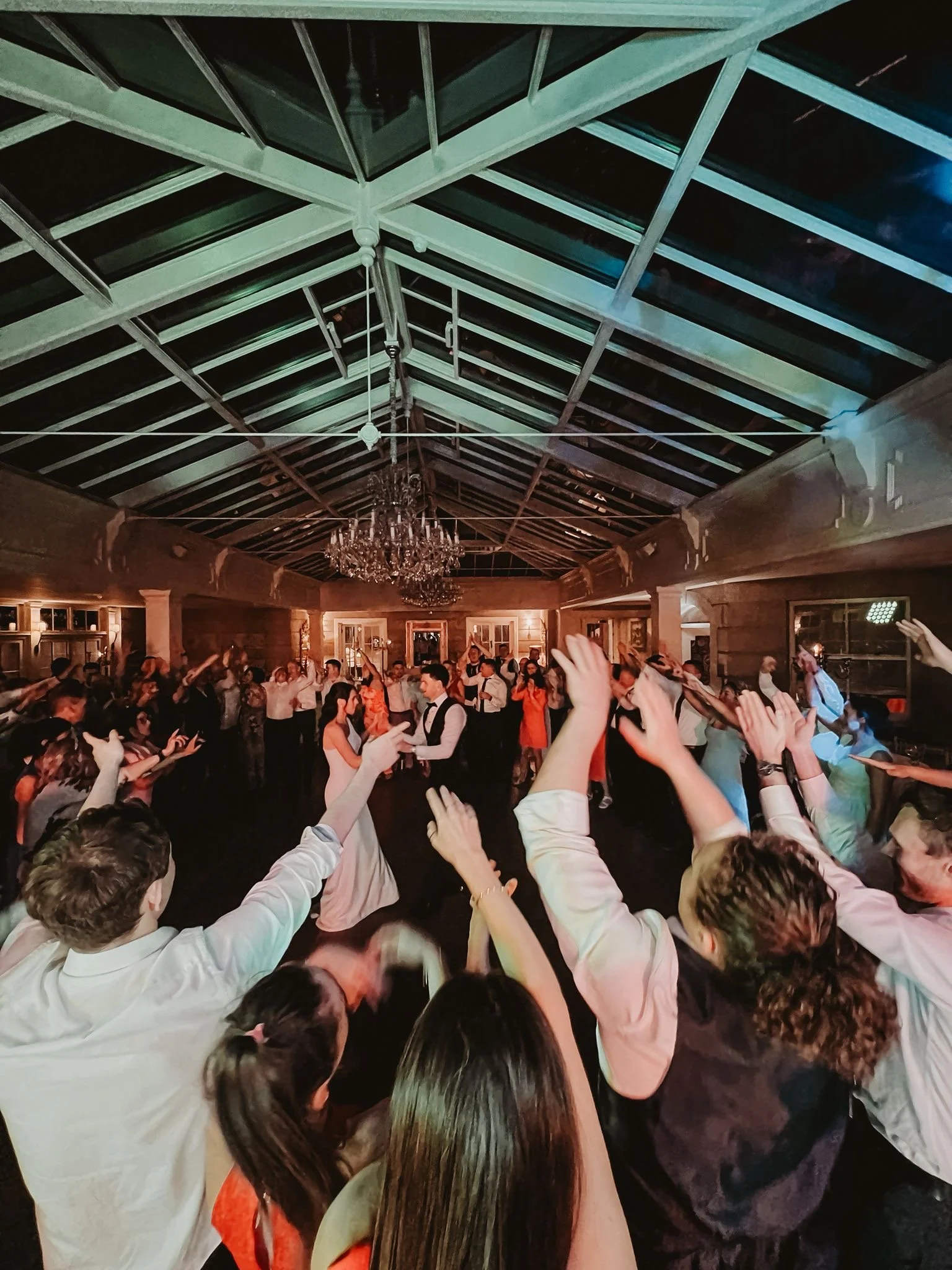 Crowd of people dancing and celebrating at a wedding reception in an indoor venue with a high ceiling, chandeliers, and wood-paneled walls.