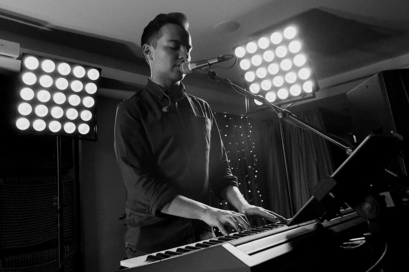Gavin Coughlin playing a keyboard with a microphone in front of him in a dimly lit room, with stage lights in the background.