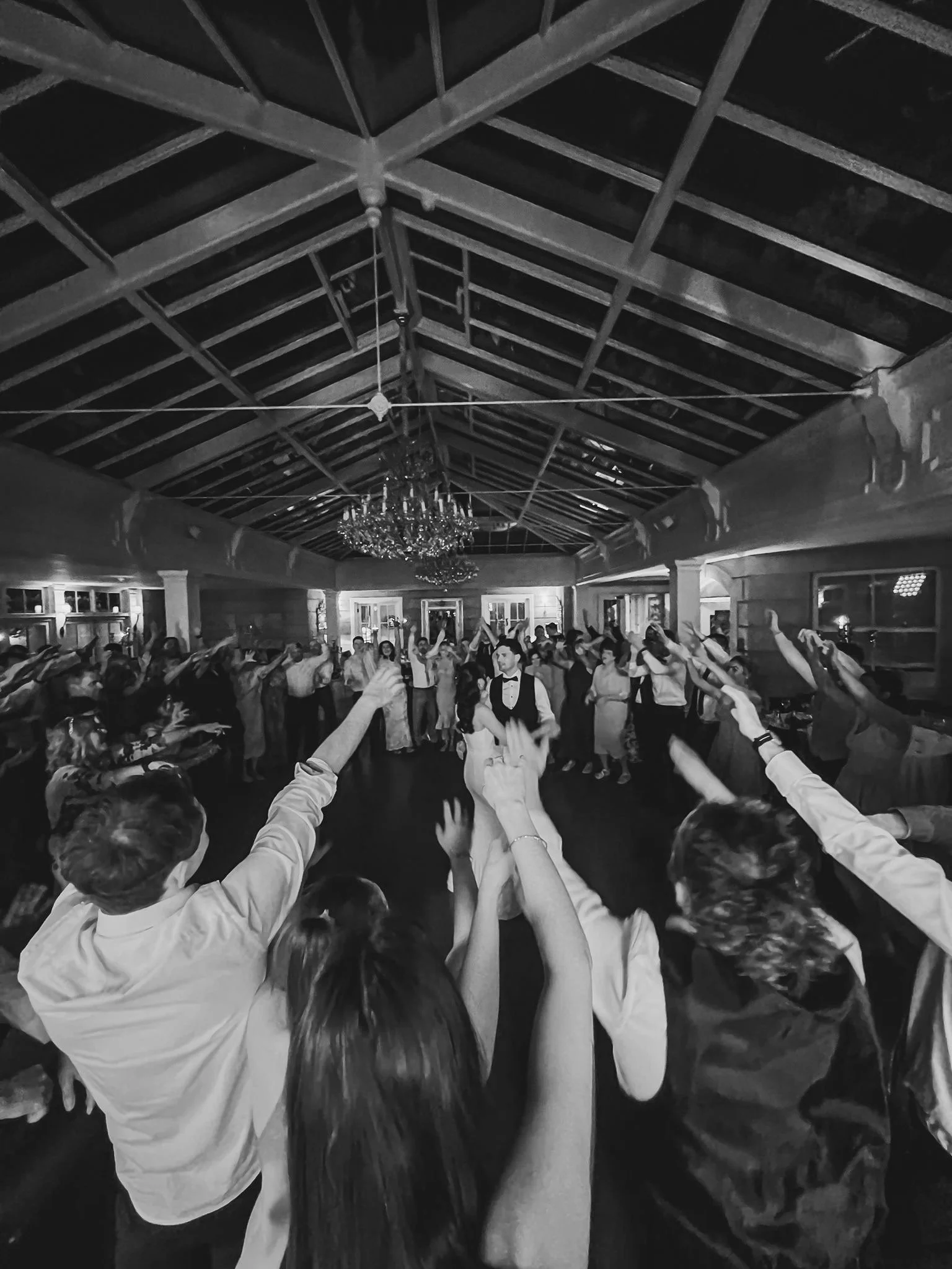 A large group of people at a Traffic Wedding Band wedding reception in Ireland, dancing with their arms raised, in a room with a high, wooden ceiling and chandeliers.