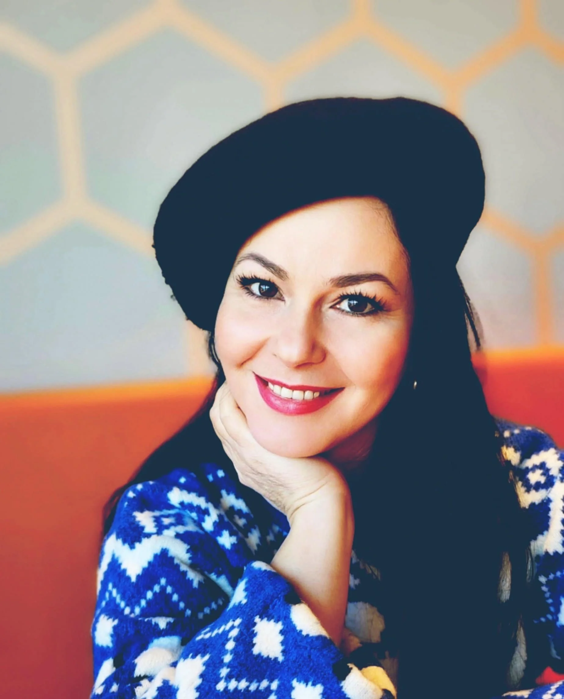A woman with black hair, smiling, wearing a black beret and a blue and white patterned sweater, resting her chin on her hand, in front of a gray background with orange accents.