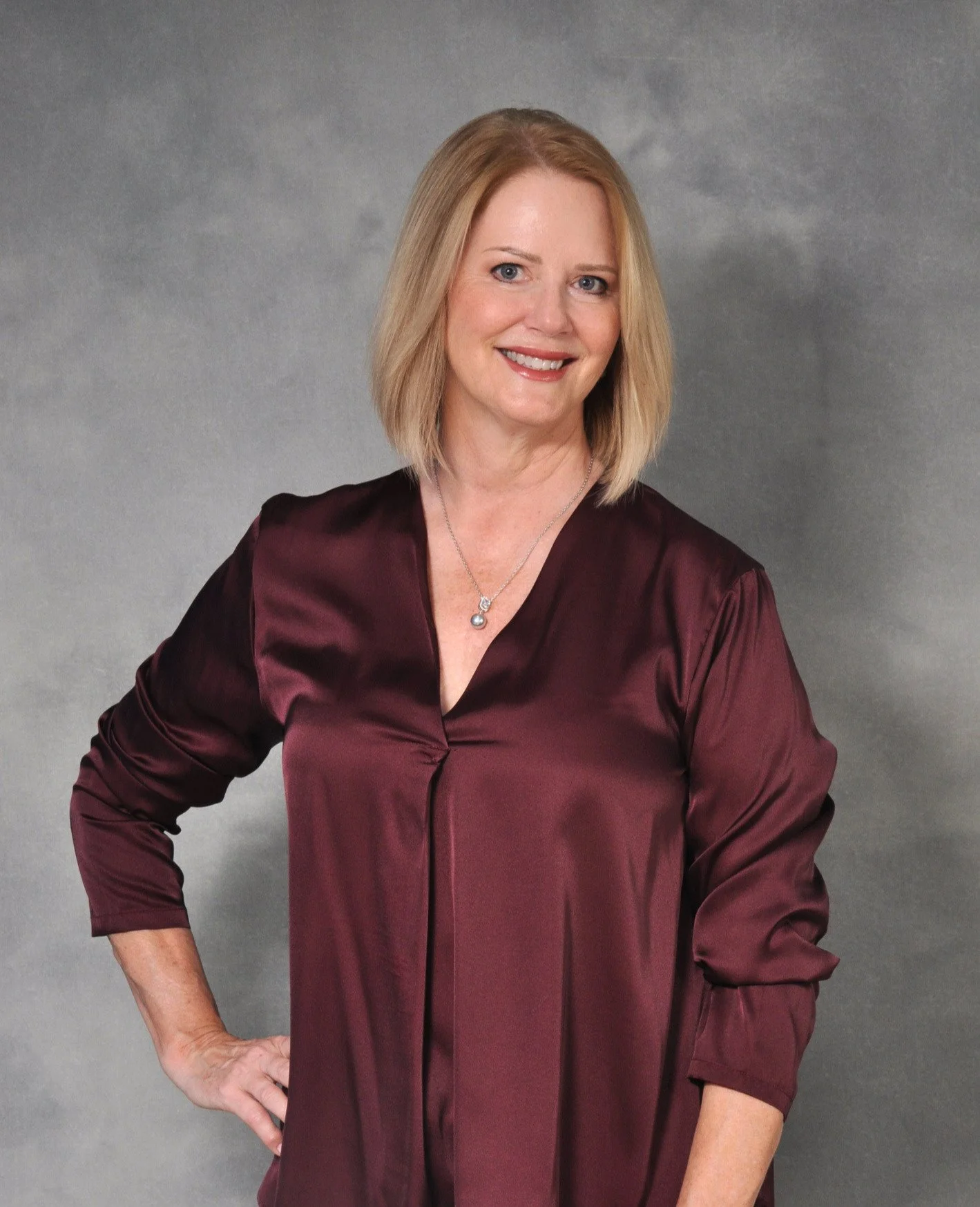 A woman with blonde hair smiling, wearing a maroon satin blouse and a silver necklace, standing against a gray backdrop.