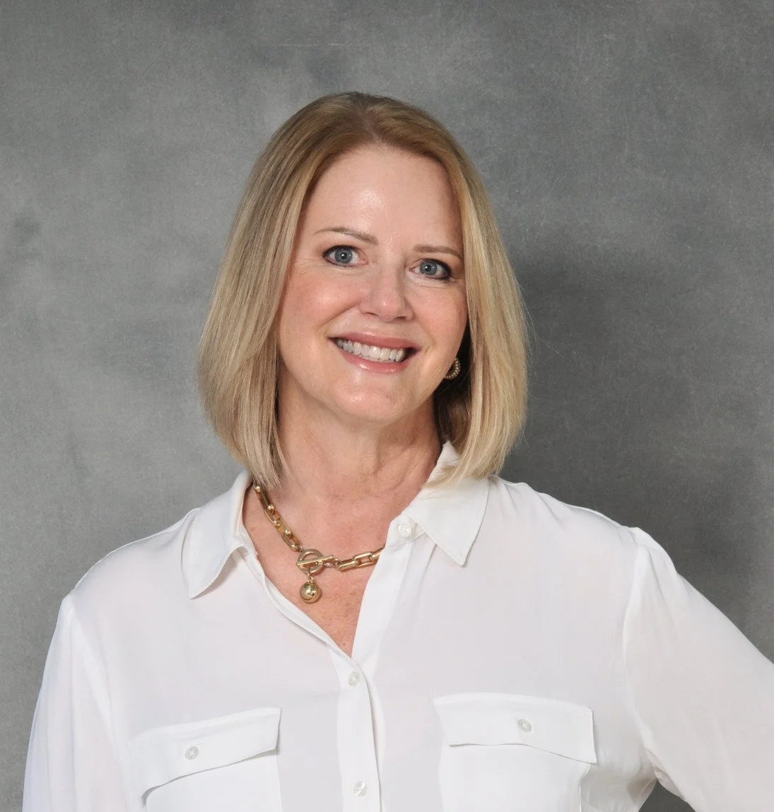 A smiling woman with shoulder-length blonde hair wearing a white button-up shirt and a gold necklace, standing against a gray background.