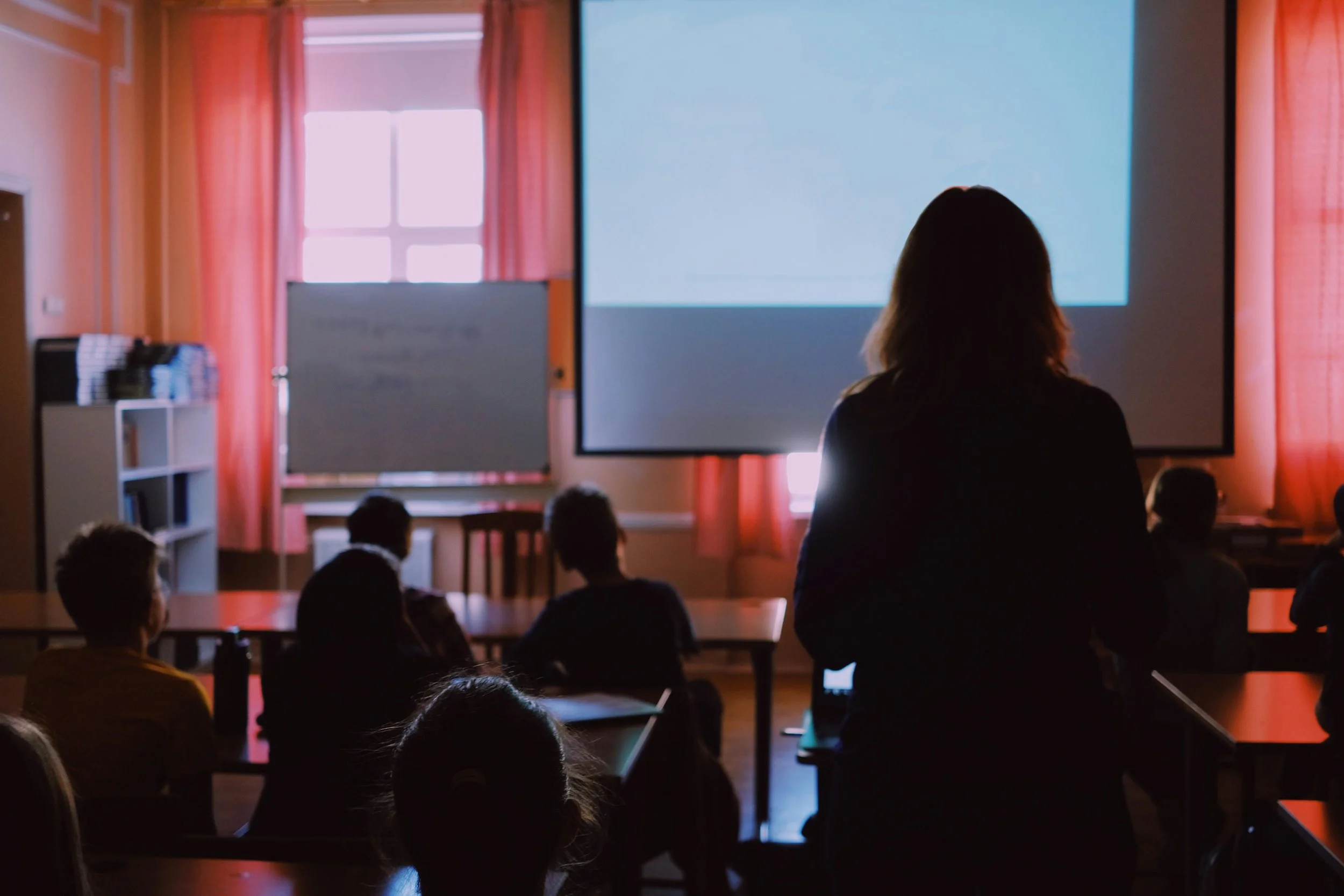 A person is standing and giving a presentation in front of a projected screen to a seated audience in a classroom or conference room.