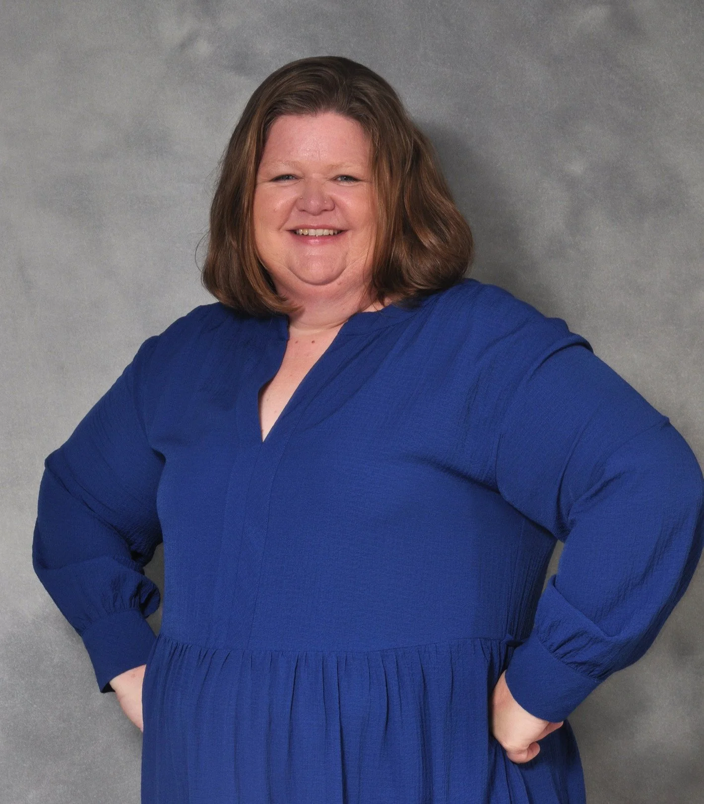 Smiling woman with shoulder-length brown hair wearing a royal blue dress with long puffed sleeves standing against a gray background.
