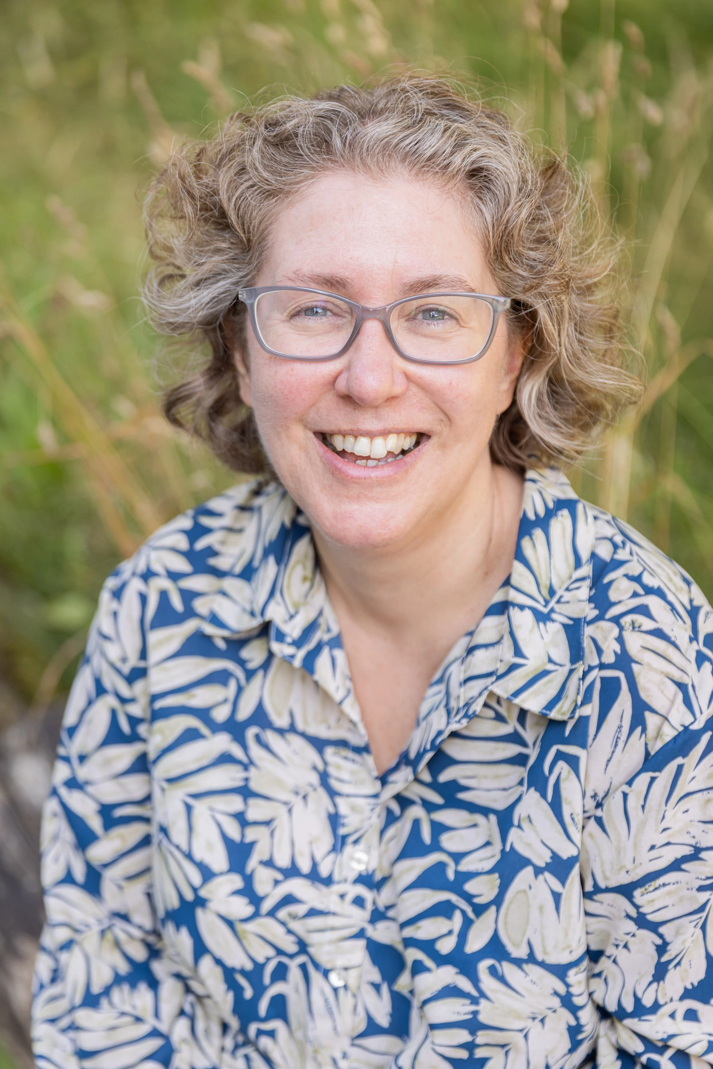 Laura Machin, a woman with curly gray hair and glasses, is smiling outdoors in front of green grass or foliage, wearing a patterned shirt.