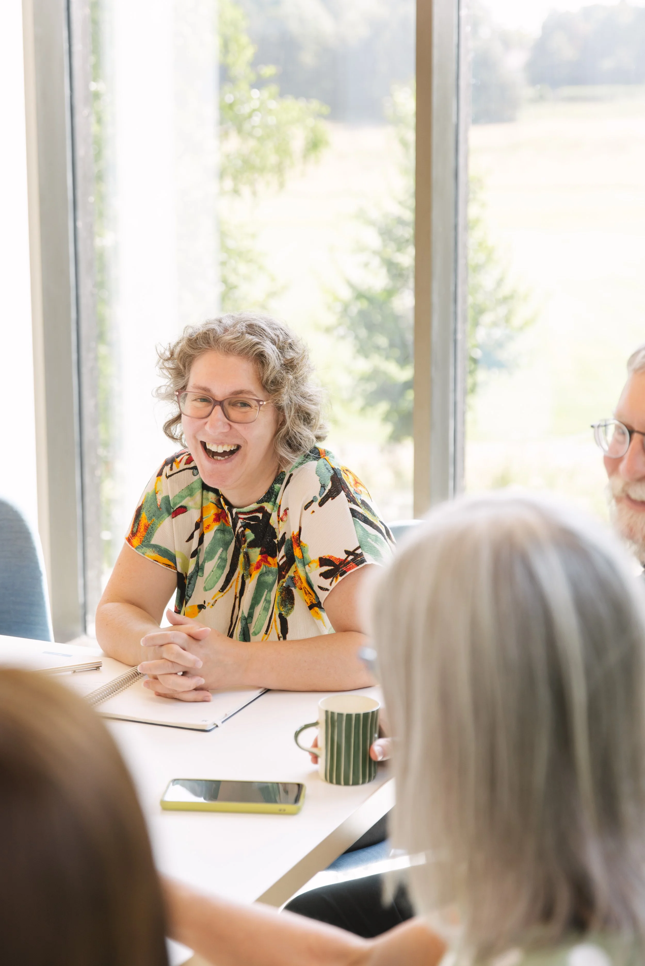 Woman with curly gray hair and glasses smiling while sitting at a table with others during a meeting, with a large window showing greenery outside.