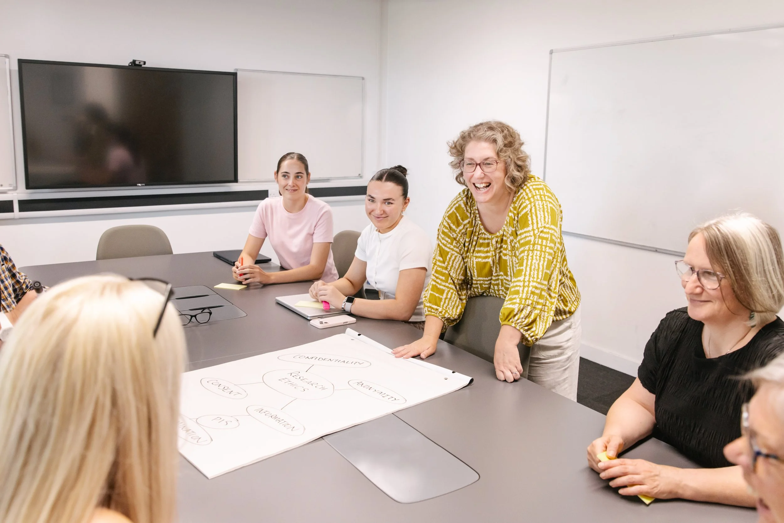 A group of women seated around a conference table in a classroom or meeting room, engaged in a discussion. One woman, standing with hands resting on the table, is smiling and talking to the group. Others are listening attentively, with notepads and writing utensils in front of them. A large whiteboard and a screen are on the wall behind them.