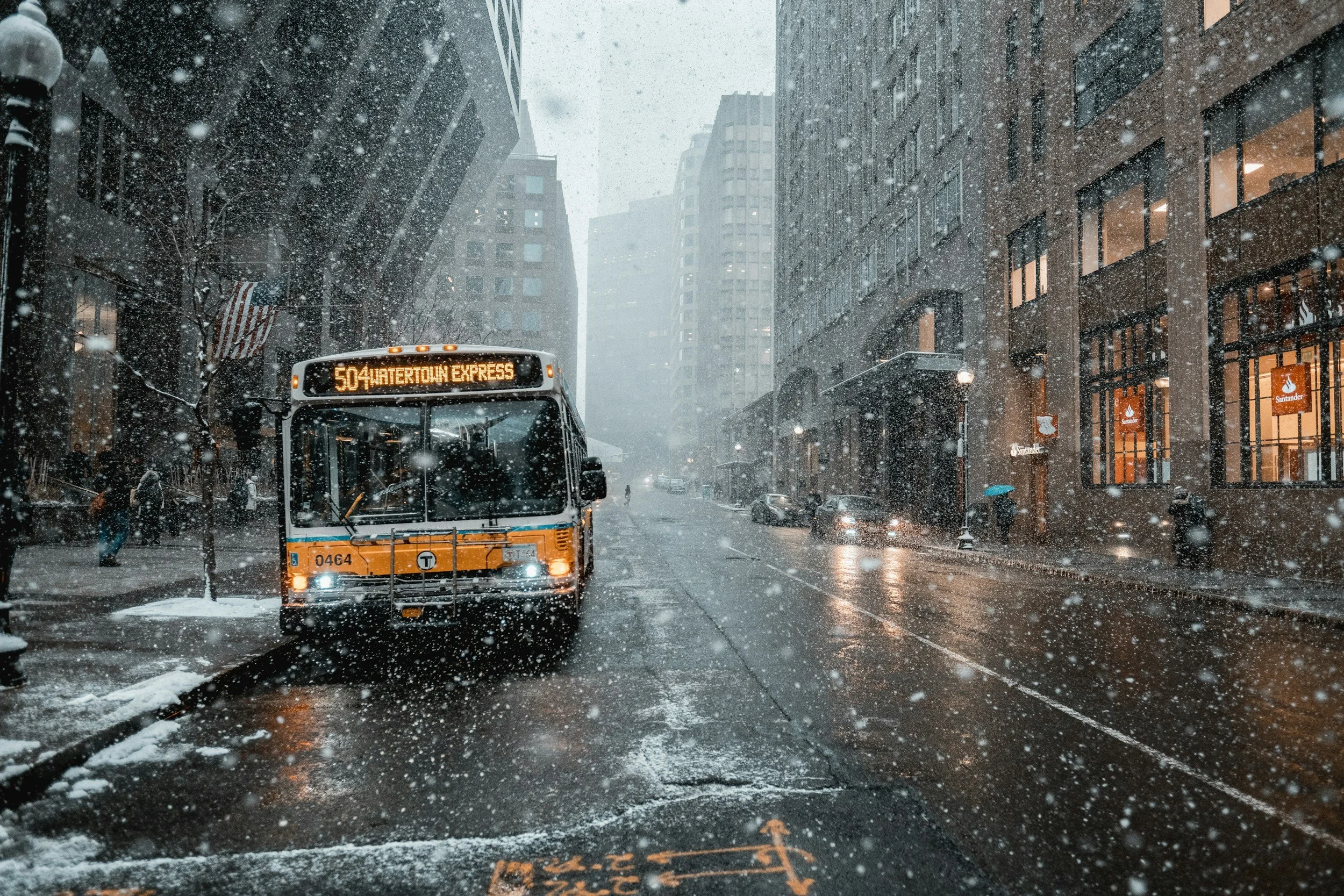 A city street with tall buildings on either side during a snowstorm. A bus with the sign '504 Watertown Express' is driving through, with snow falling heavily and accumulating on the street and vehicles.