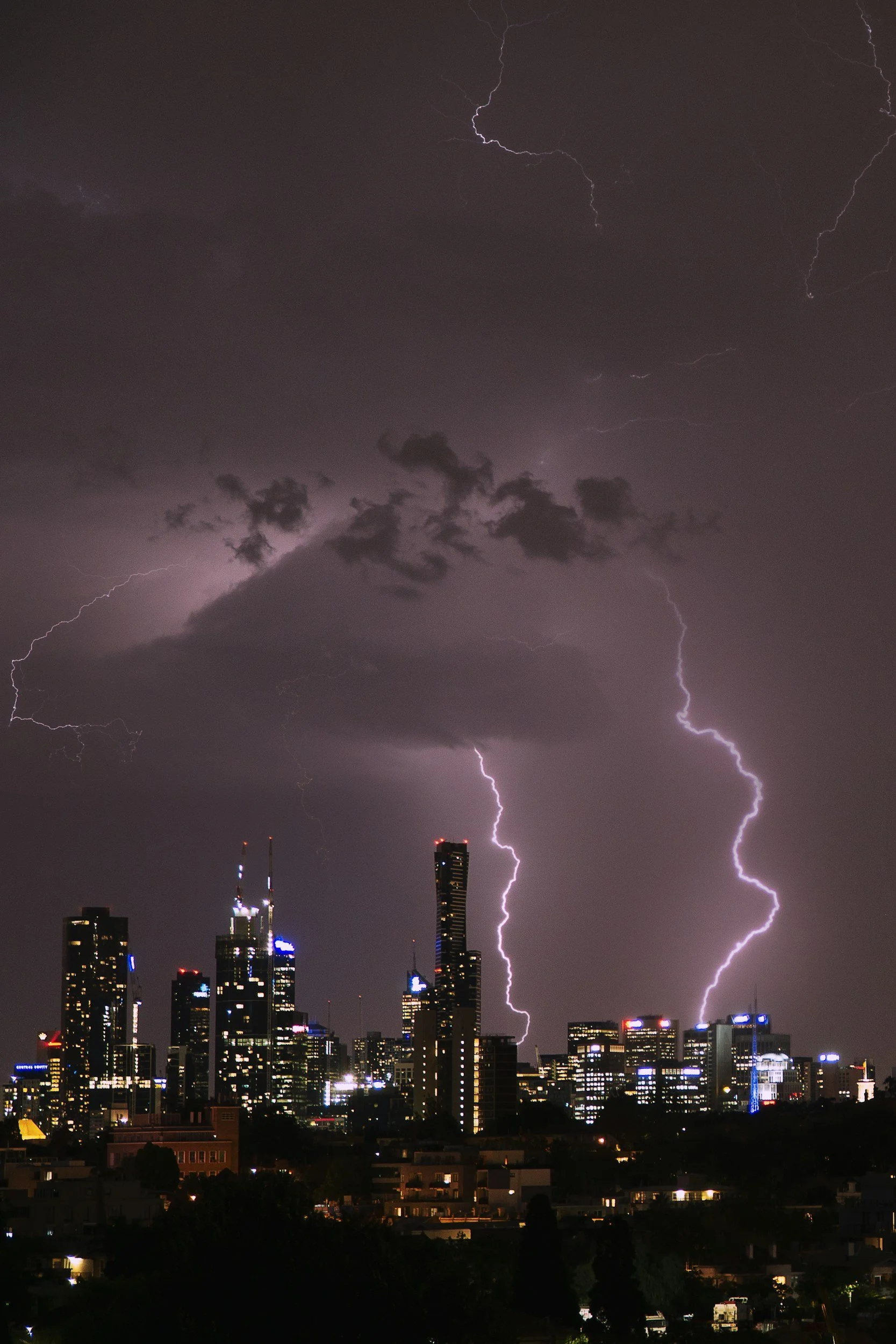 Nighttime city skyline with multiple lightning bolts striking down from dark, stormy clouds.
