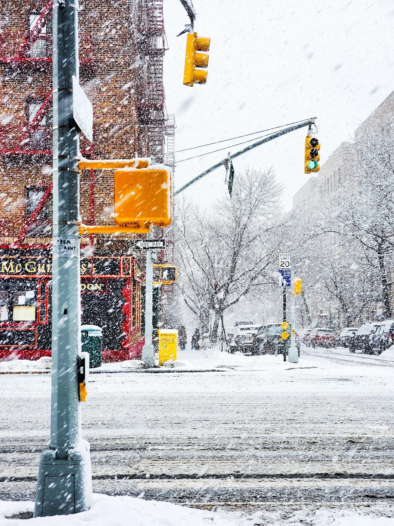 Snow-covered city street with traffic signals, a brick building, trees, parked cars, and snow falling heavily.