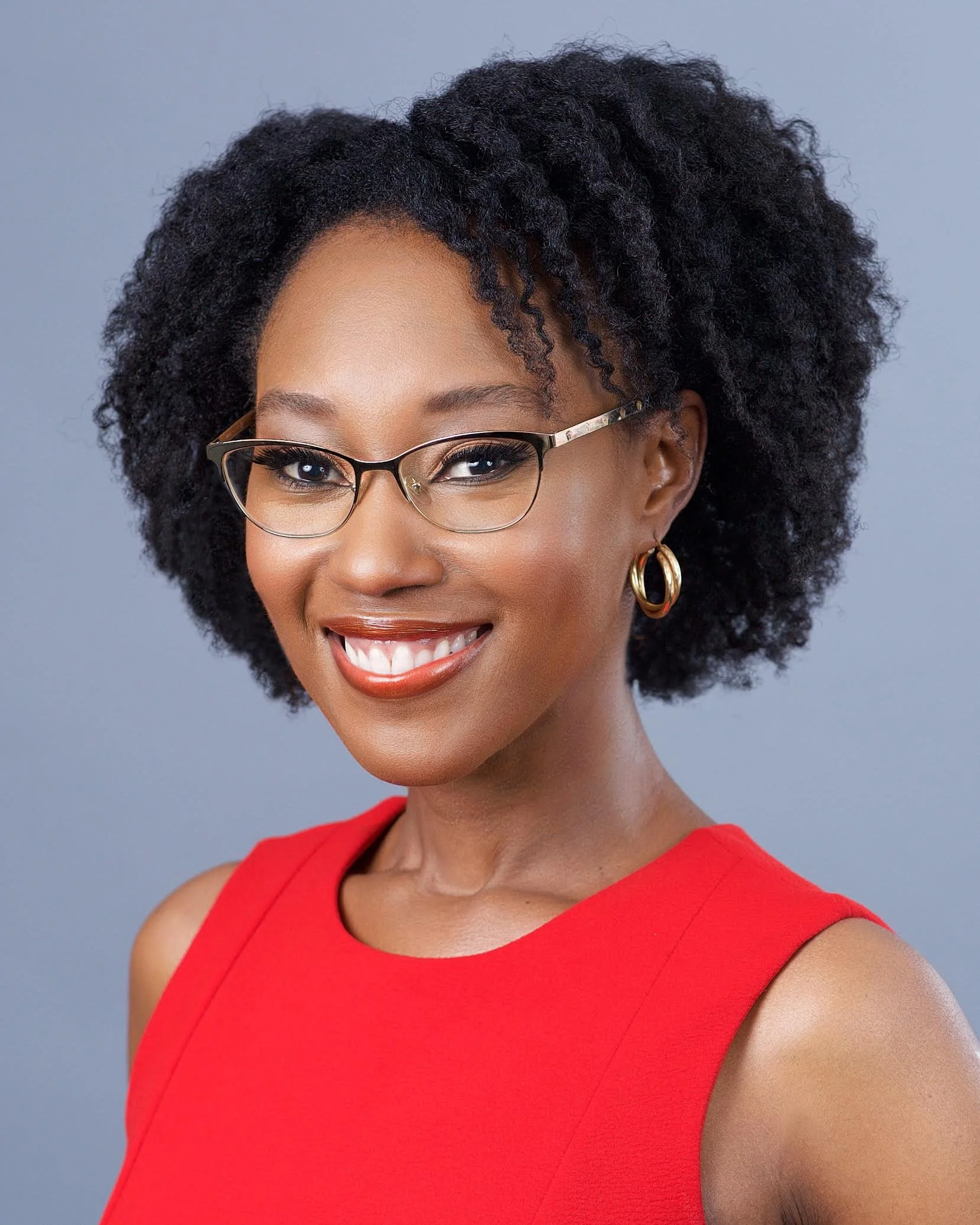 Close-up of a smiling African American woman wearing stylish glasses, gold hoop earrings, and a red sleeveless dress, with a short, curly hairstyle against a plain gray background.