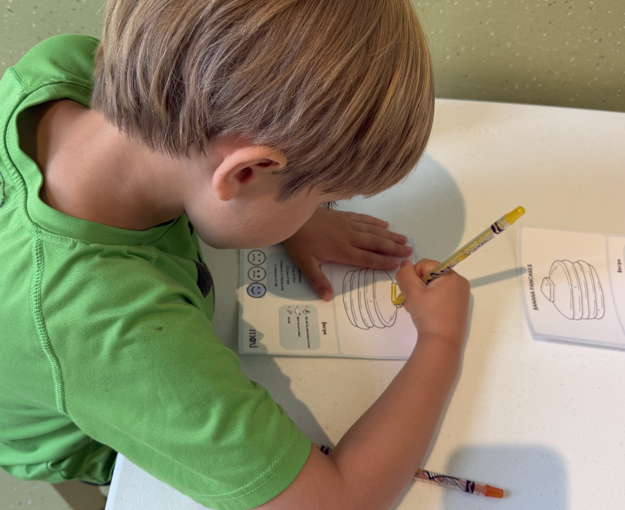 A young boy in a green shirt coloring a workbook at a white table.