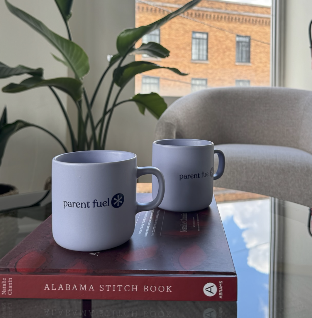 Two white coffee mugs with 'parent fuel' text on a glass table, with a large green plant, an armchair, and a large window in the background.