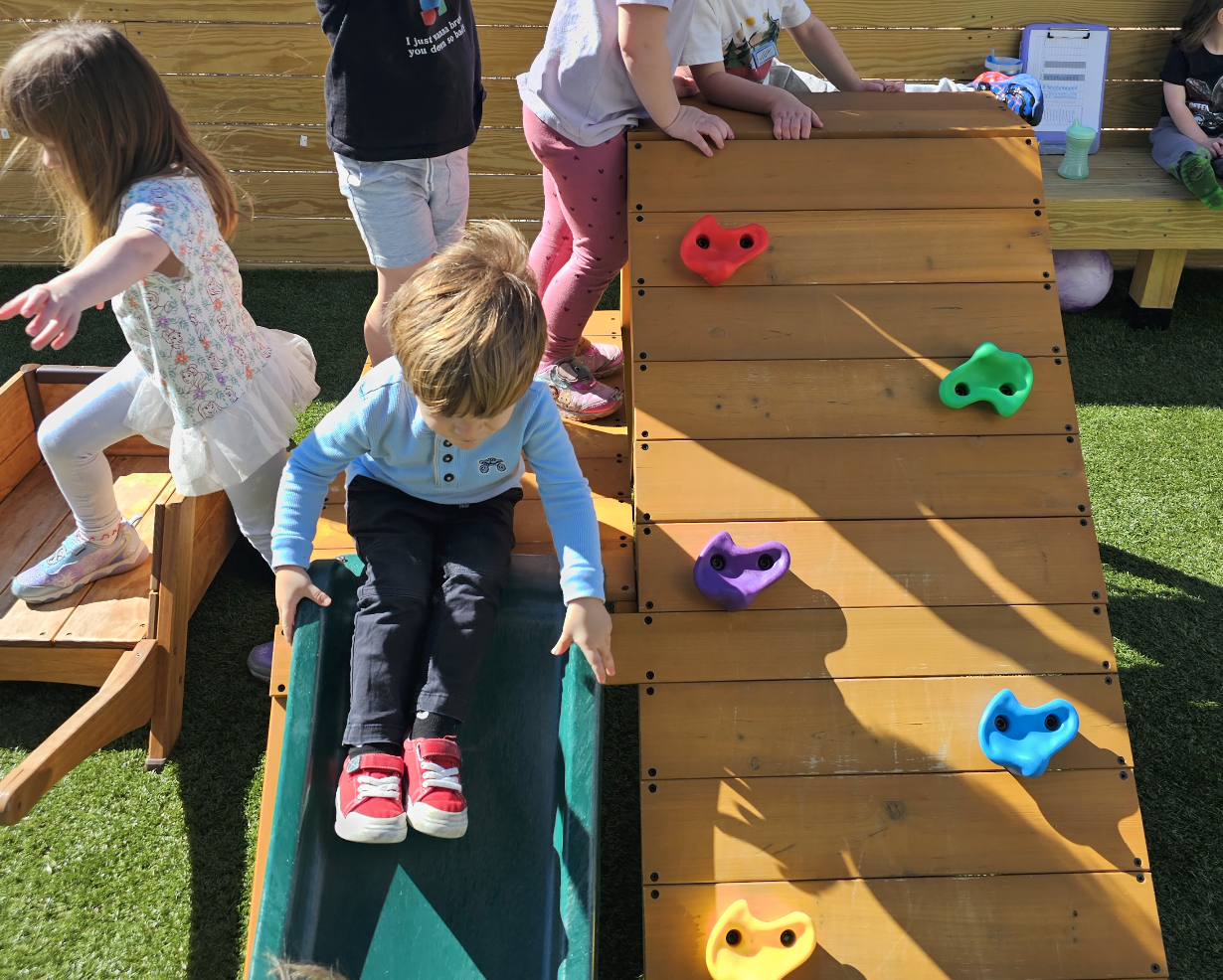 Children playing on a wooden playground structure with a slide and climbing wall with colorful holds.