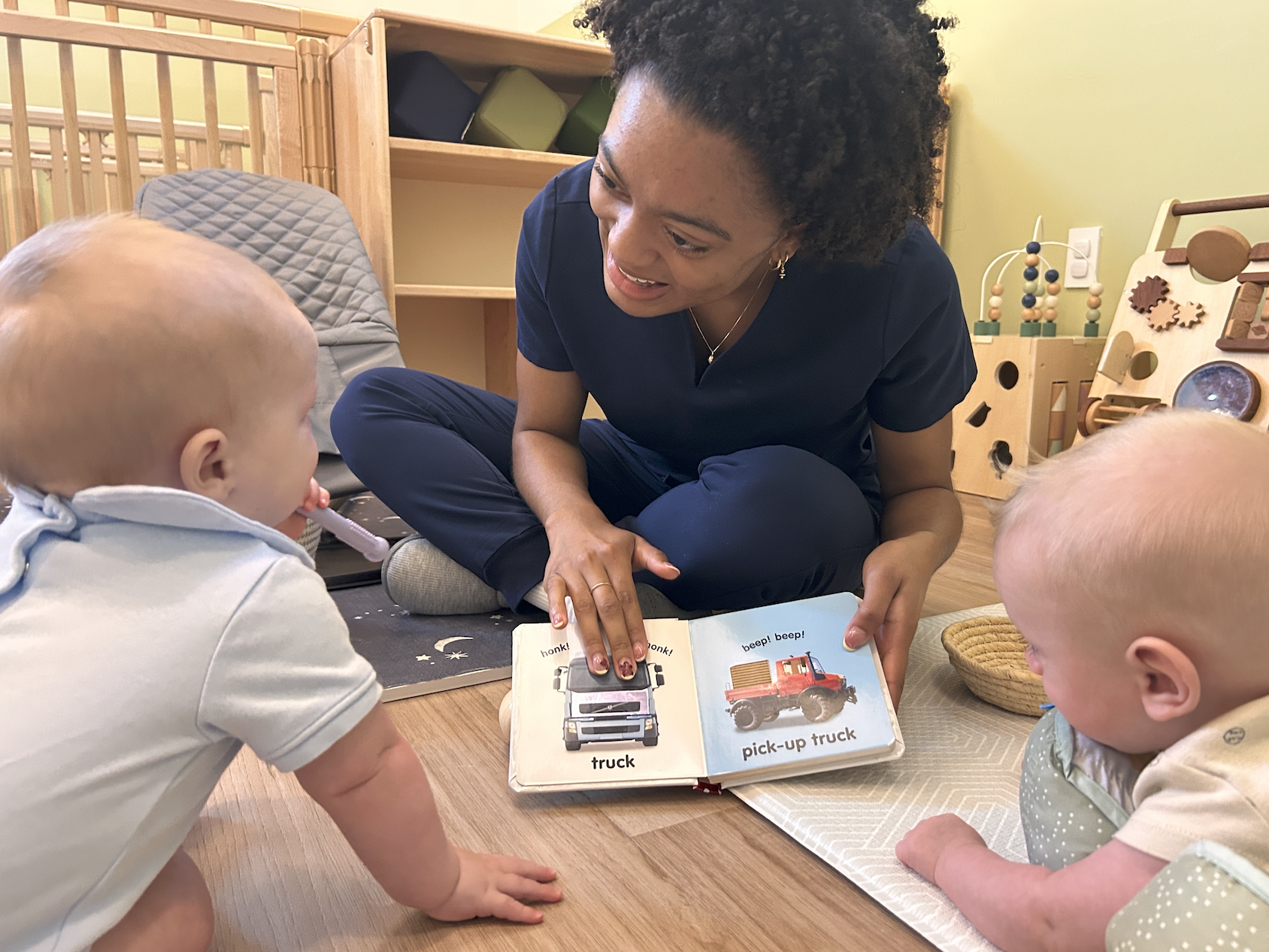 A teacher reading a children's book to two young infants in a playroom with wooden toys.