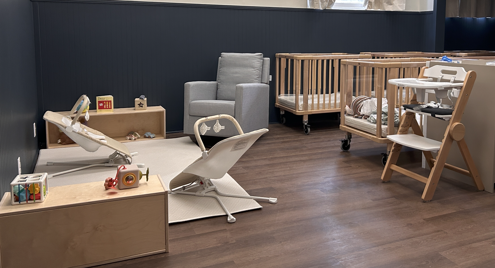 A nursery room with wooden cribs, armchair, a changing table with toys, a high chair, and a wooden shelf with toys against a blue wall and wooden flooring.